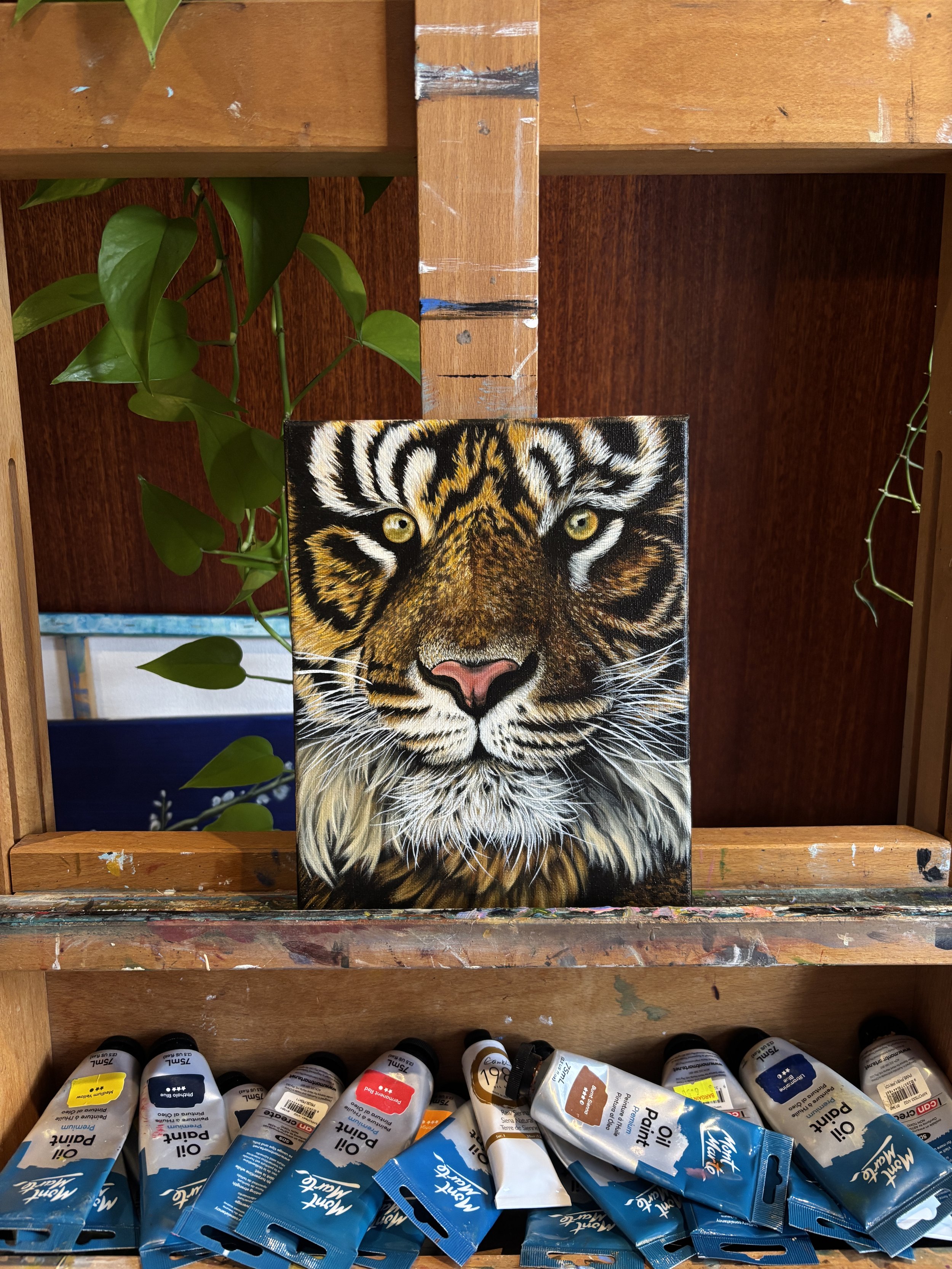 Painting of a tiger's face placed on a wooden shelf surrounded by green plants and artist's paint tubes.