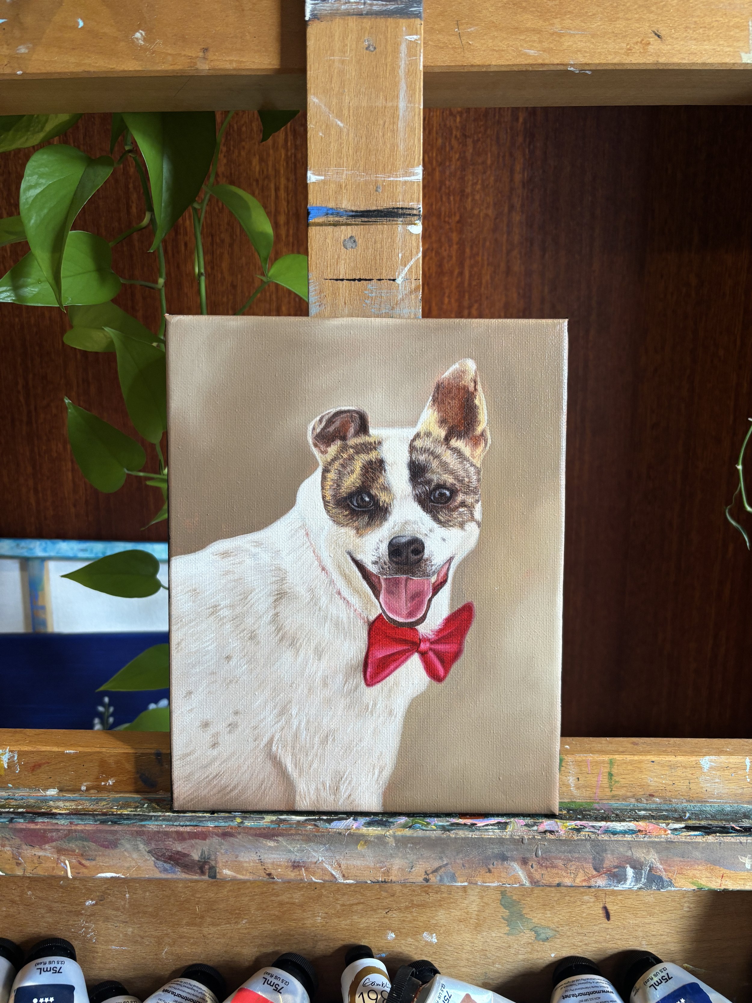Painting of a happy dog with a red bow tie, standing on a wooden art easel, surrounded by plants and art supplies.