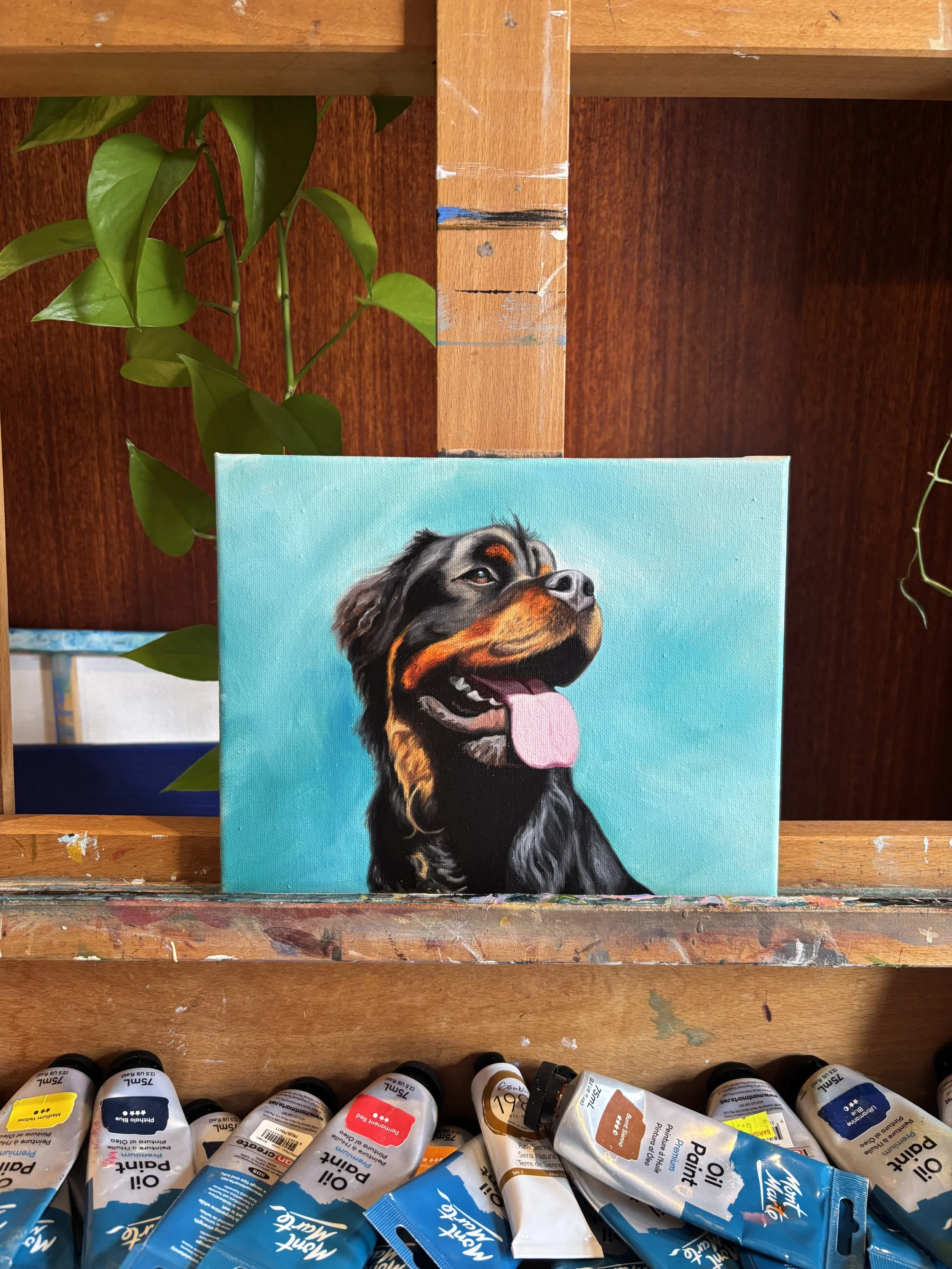 A painting of a happy Rottweiler dog with its tongue hanging out, set against a bright blue background, on an art easel surrounded by watercolor paint tubes.