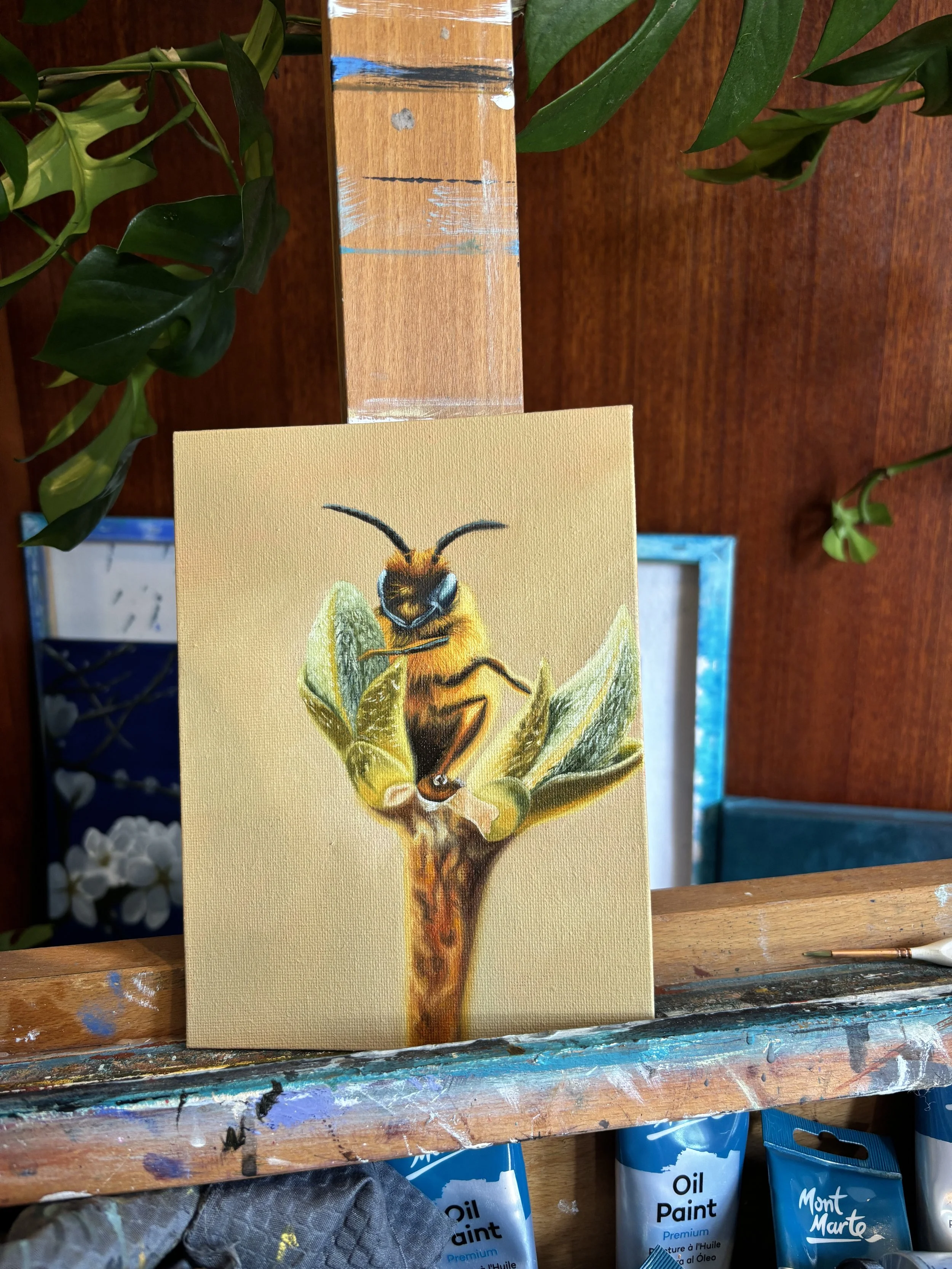 A small painting of a bee sitting on a branch with green leaves, situated on an artist's easel and surrounded by painting supplies.