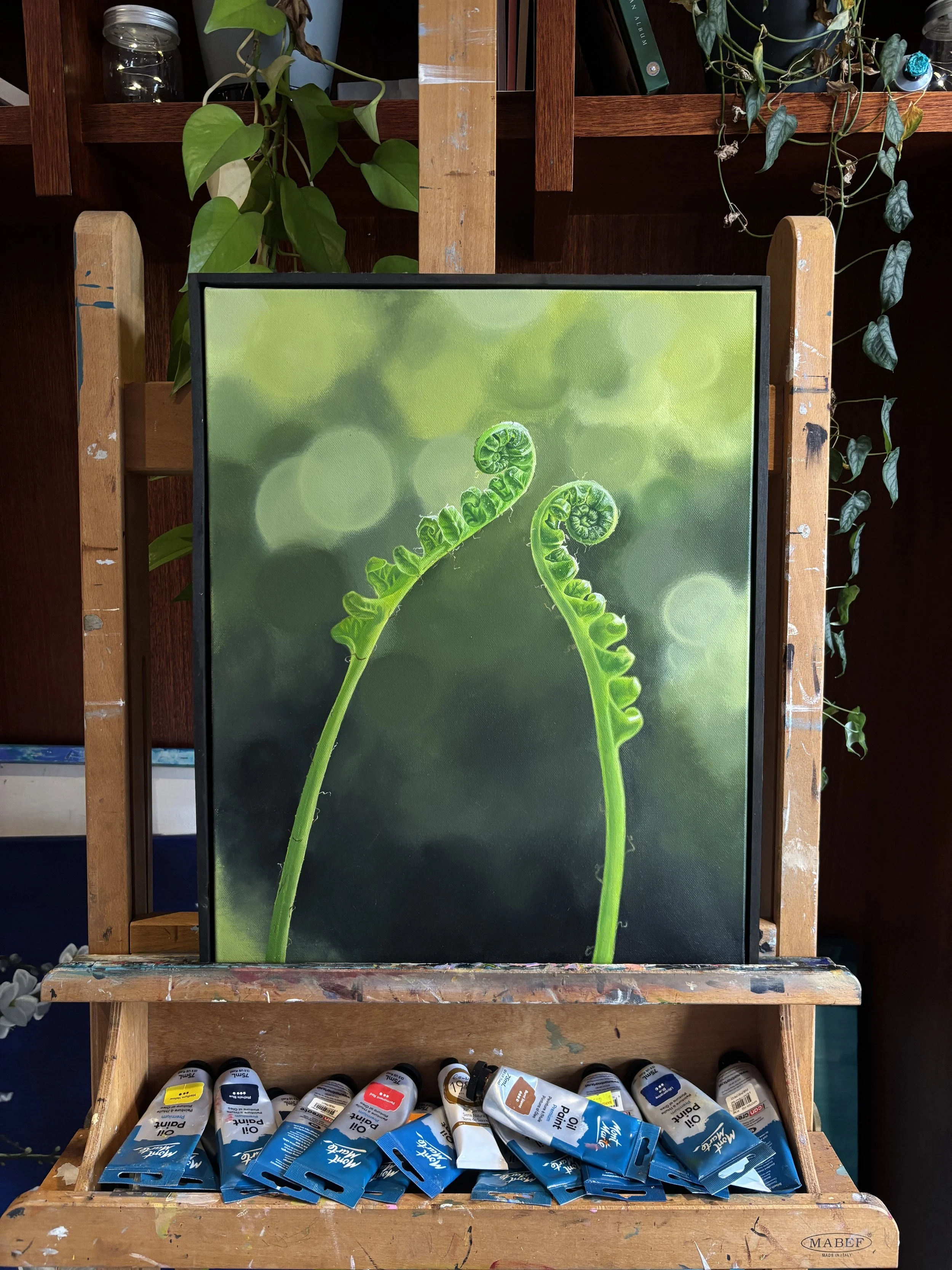 Painting of two green fern fronds unfurling, set on an easel with paint tubes at the bottom, surrounded by potted plants in a room.