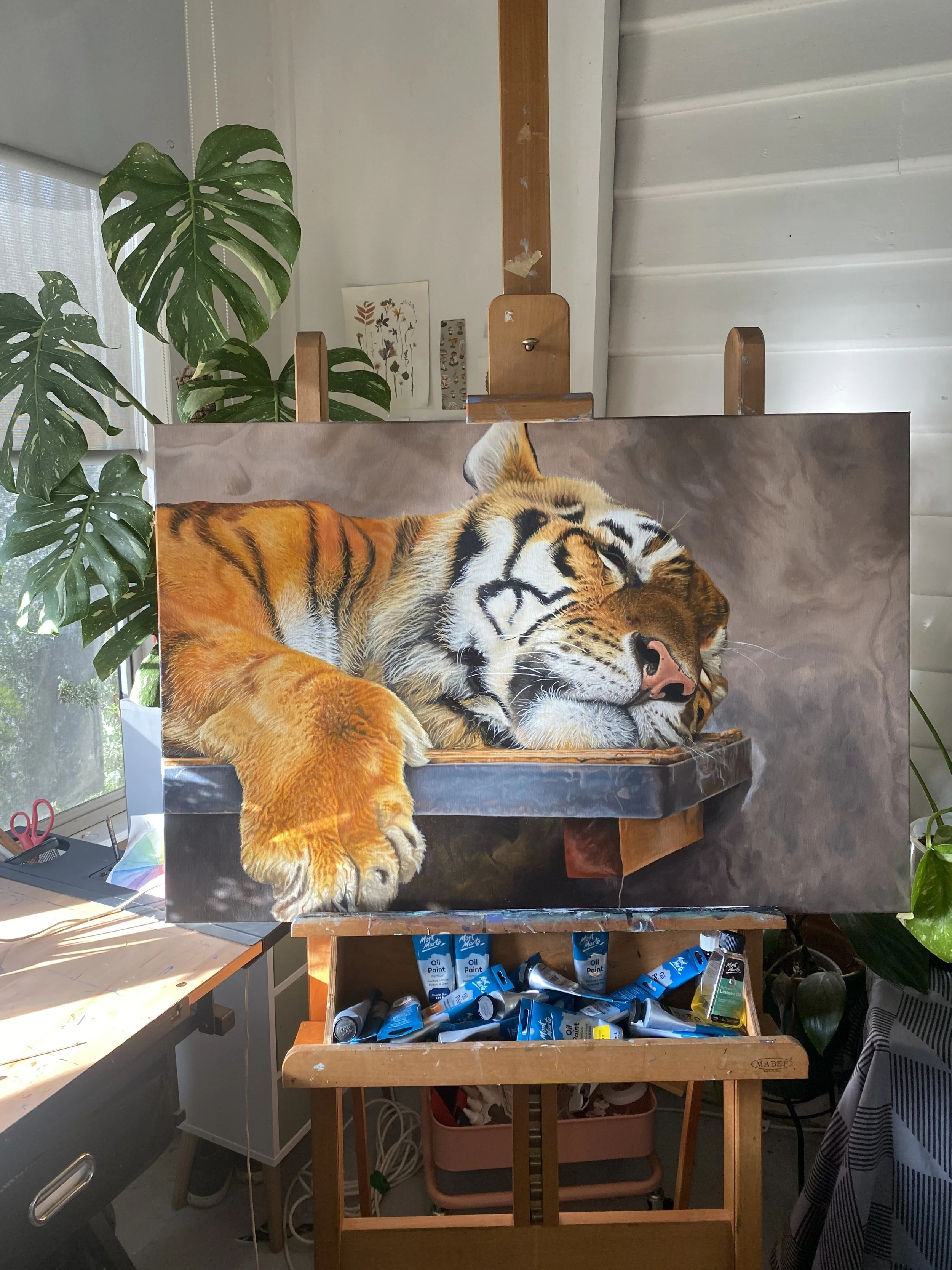 Painting of a sleeping tiger lying on a wooden platform, resting its head and front paw, set on an easel with tubes of oil paint underneath, surrounded by indoor plants.