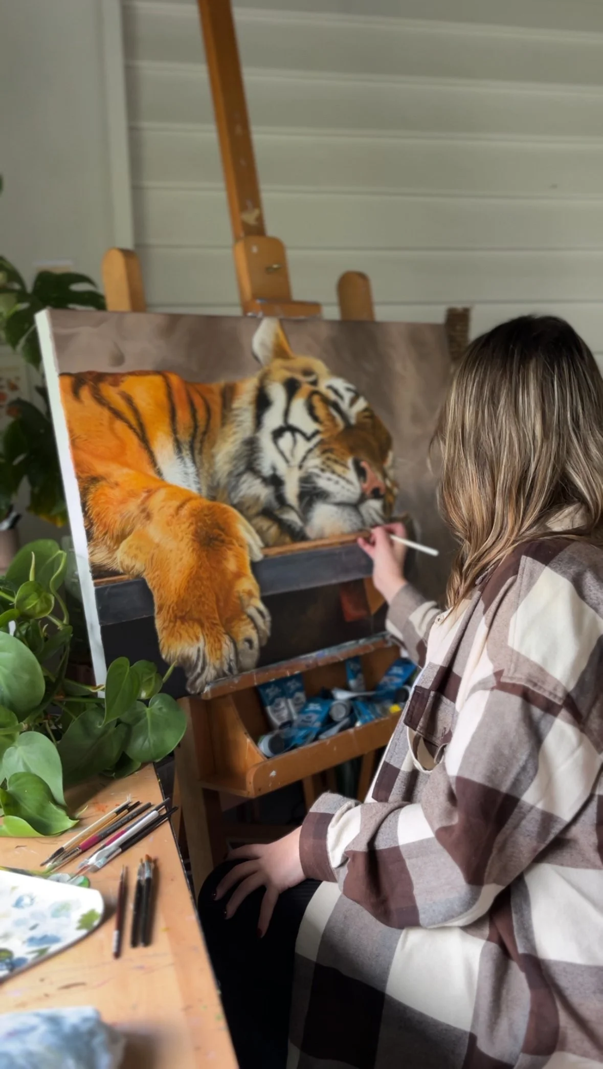 Artist painting a realistic tiger on canvas in a studio with art supplies and greenery.