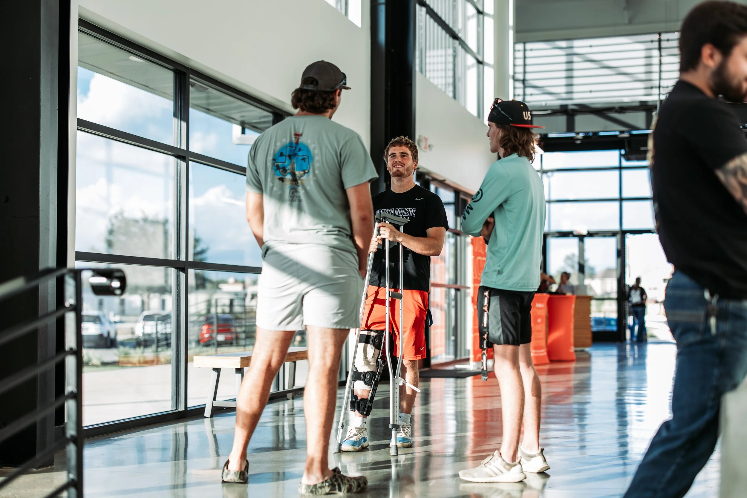 Group of young men inside a modern building, one with a leg brace talking to the others, with large windows and natural light.