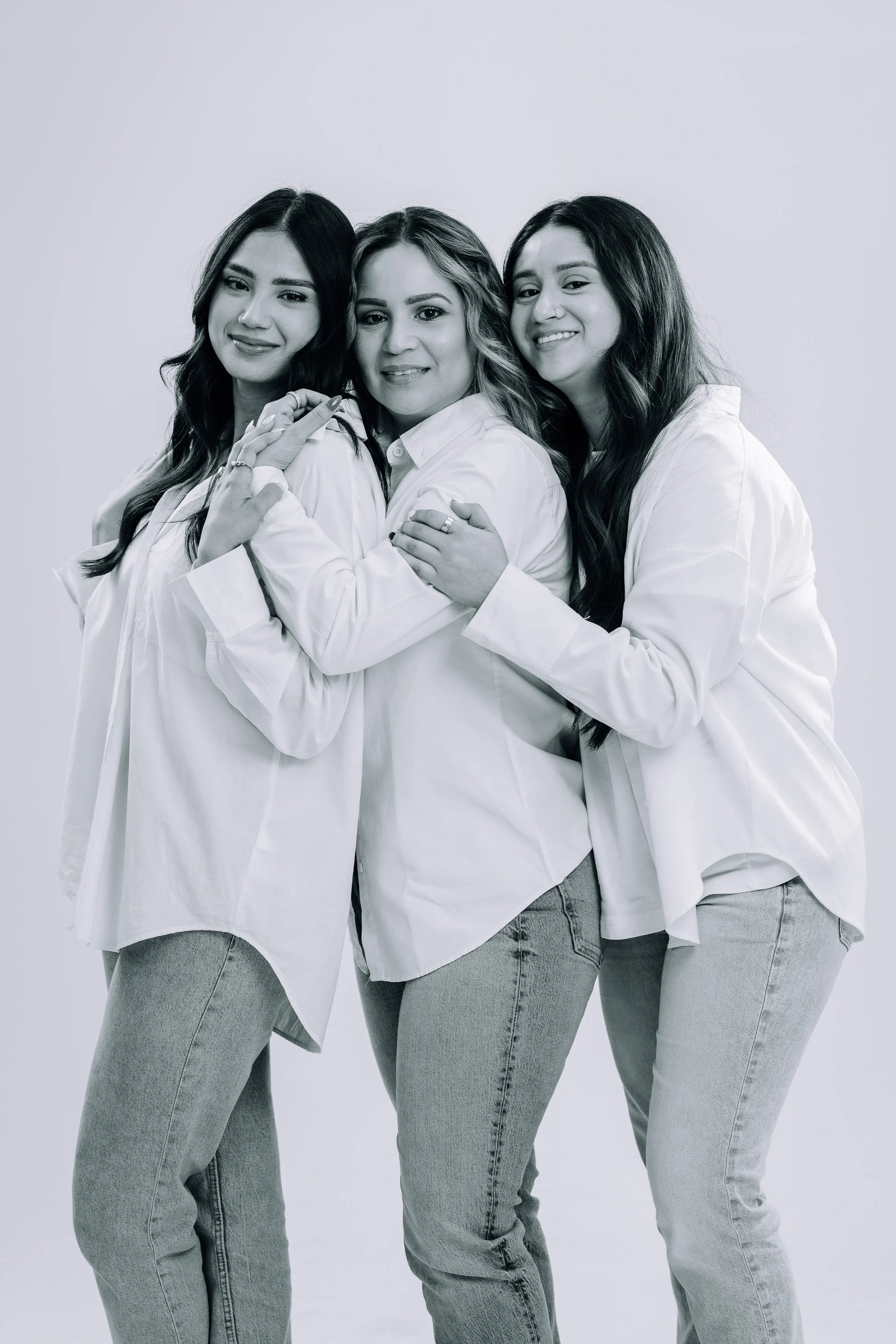 Three women in white shirts and jeans, standing together in a close embrace, smiling.
