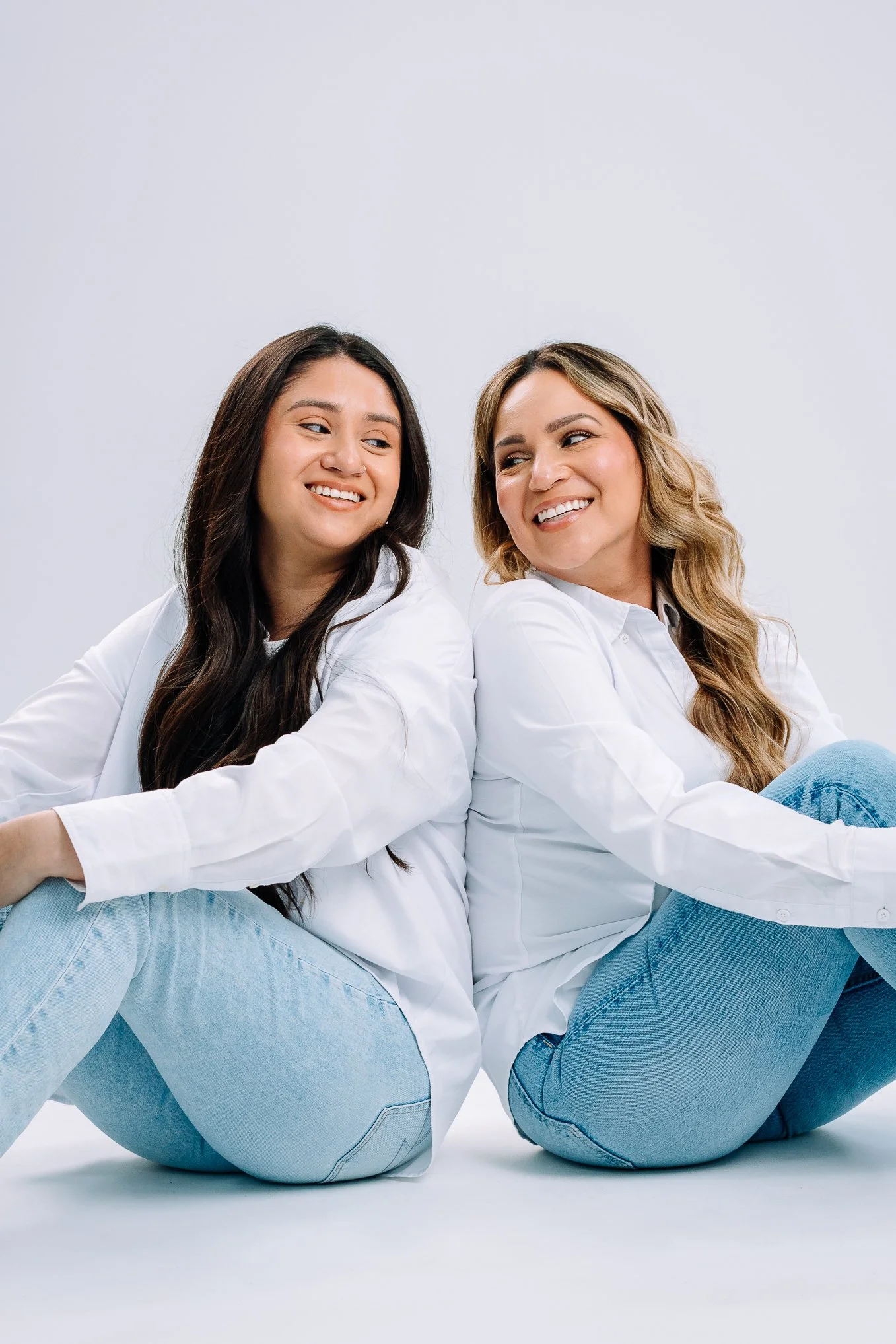 Two women sitting back-to-back, smiling and looking at each other, wearing white shirts and light blue jeans, on a white background.