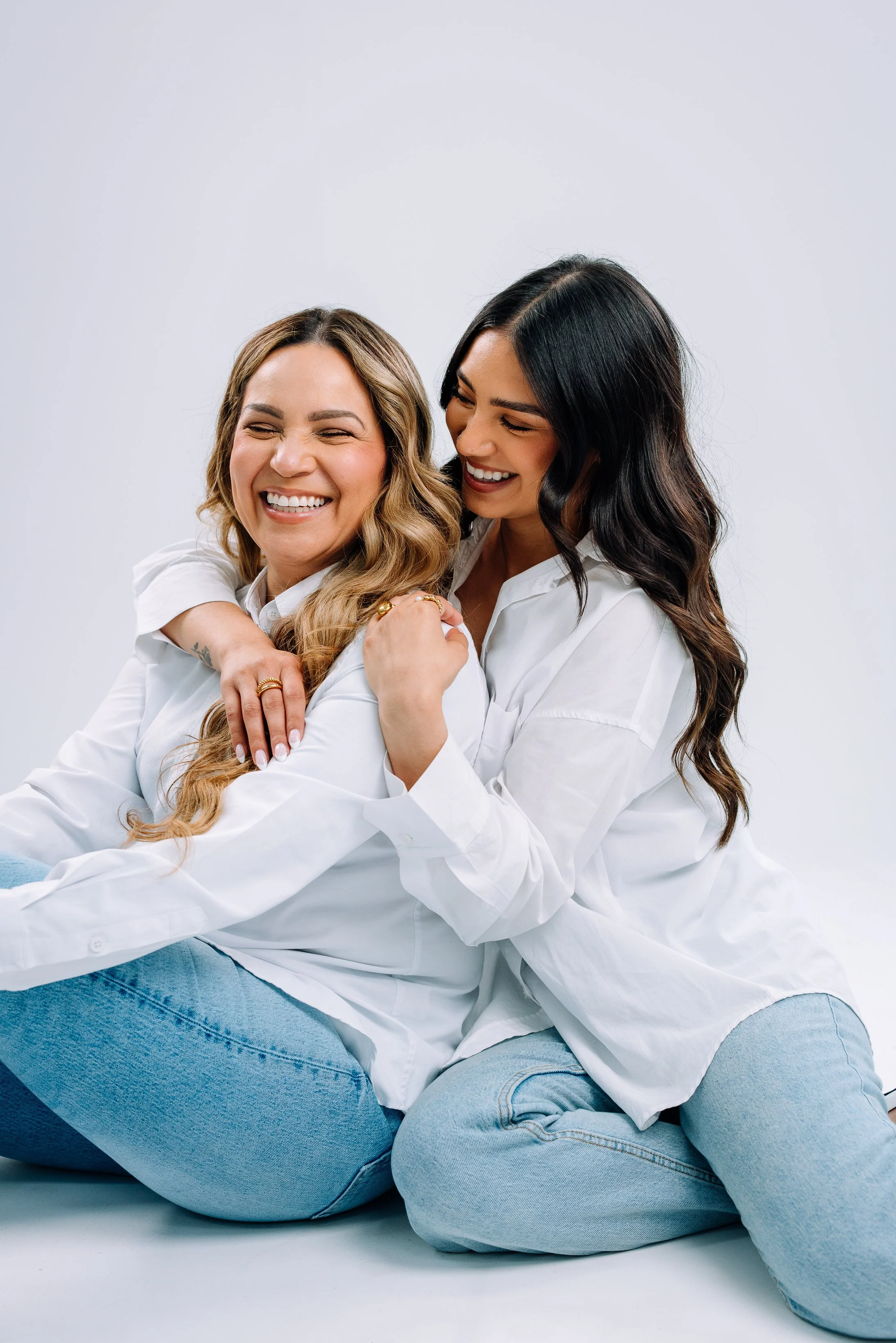 Two women sitting on the floor, smiling and hugging each other, wearing white shirts and blue jeans, against a plain white background.