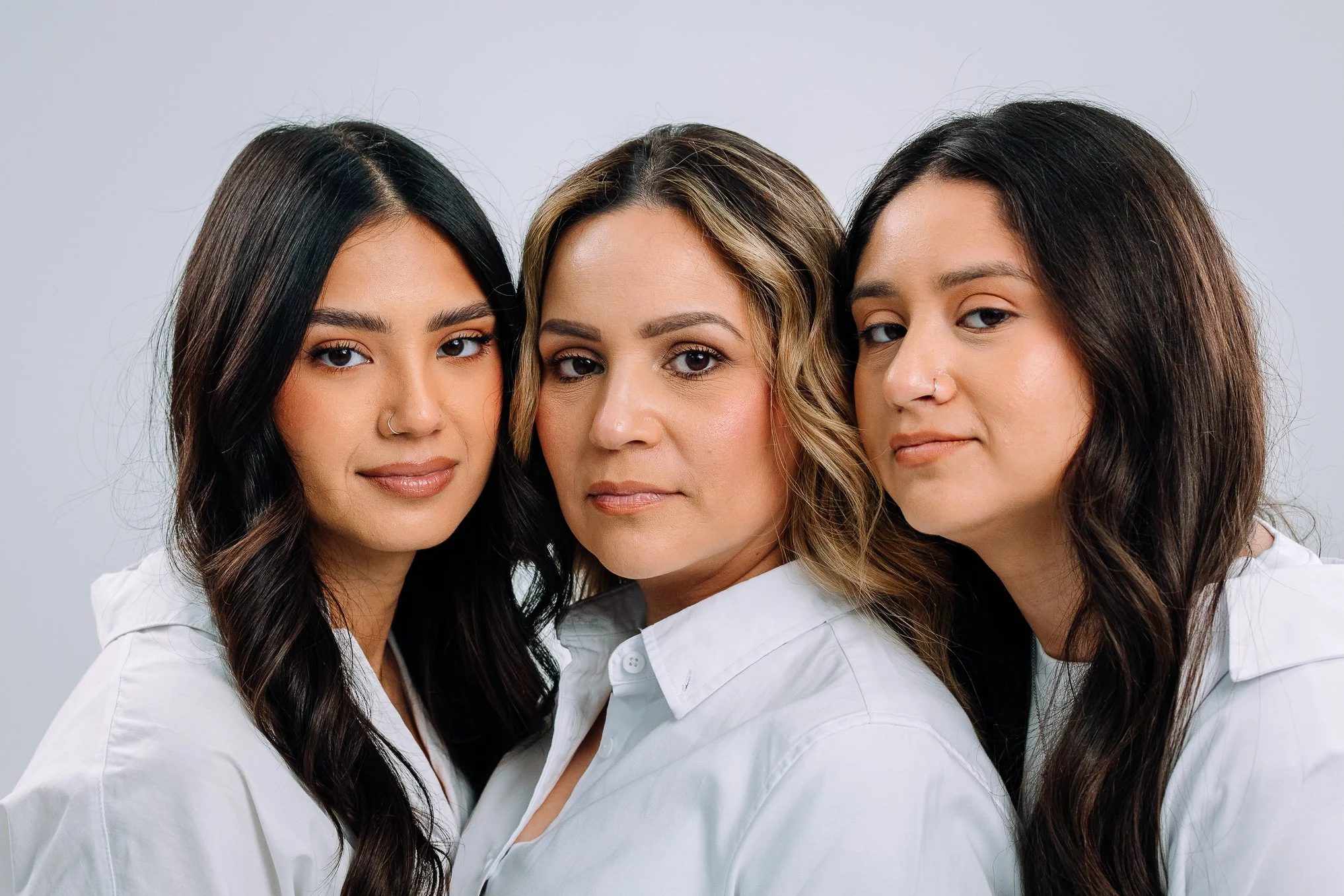 Three women with dark hair and light skin in white shirts posing closely against a plain gray background.