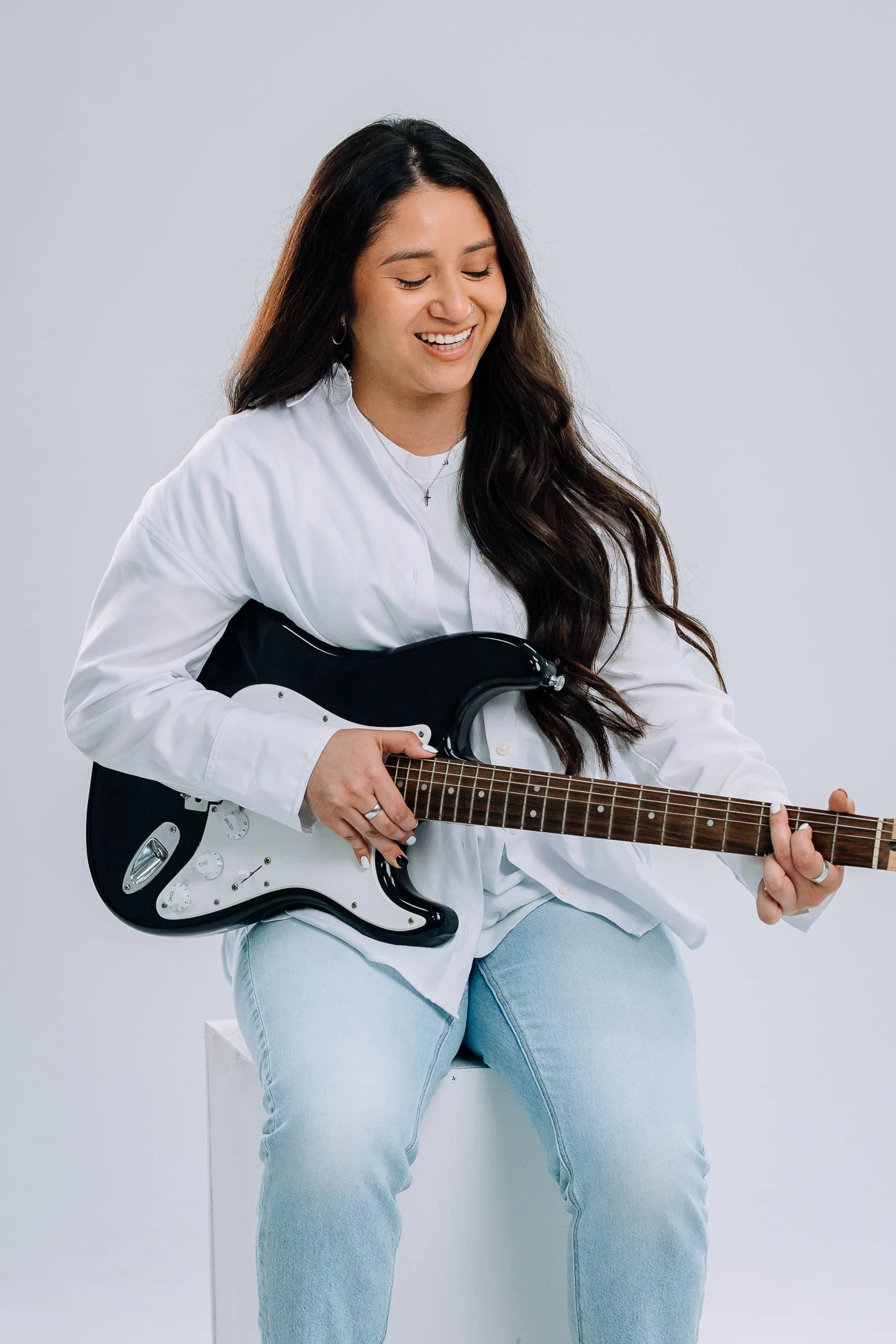 A woman with long dark hair is sitting and smiling while holding a black and white electric guitar.