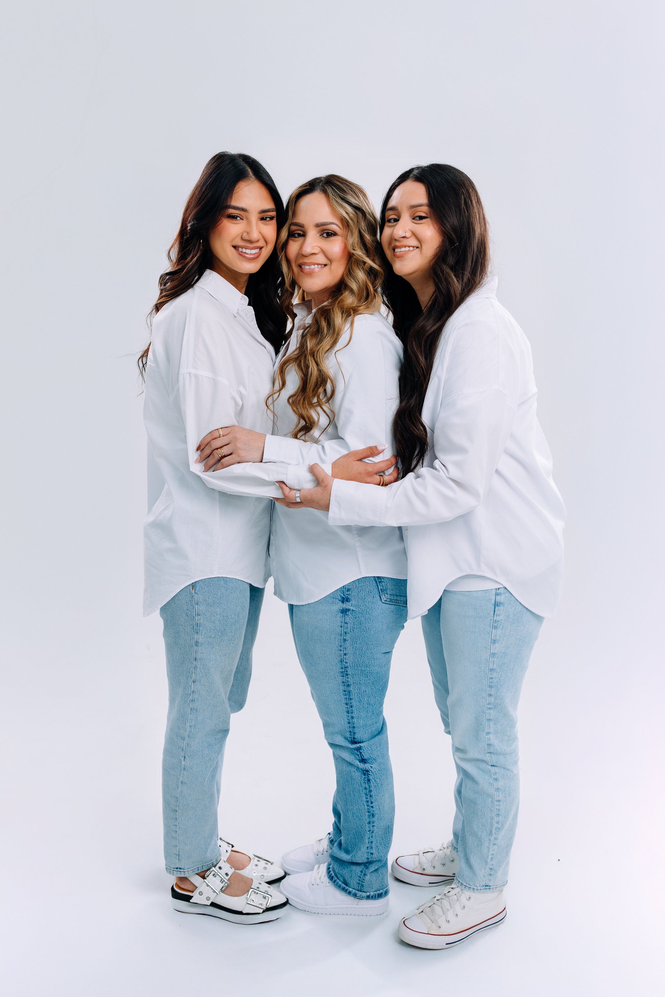 Three women standing together, hugging, dressed in white shirts and light blue jeans, smiling against a plain white background.