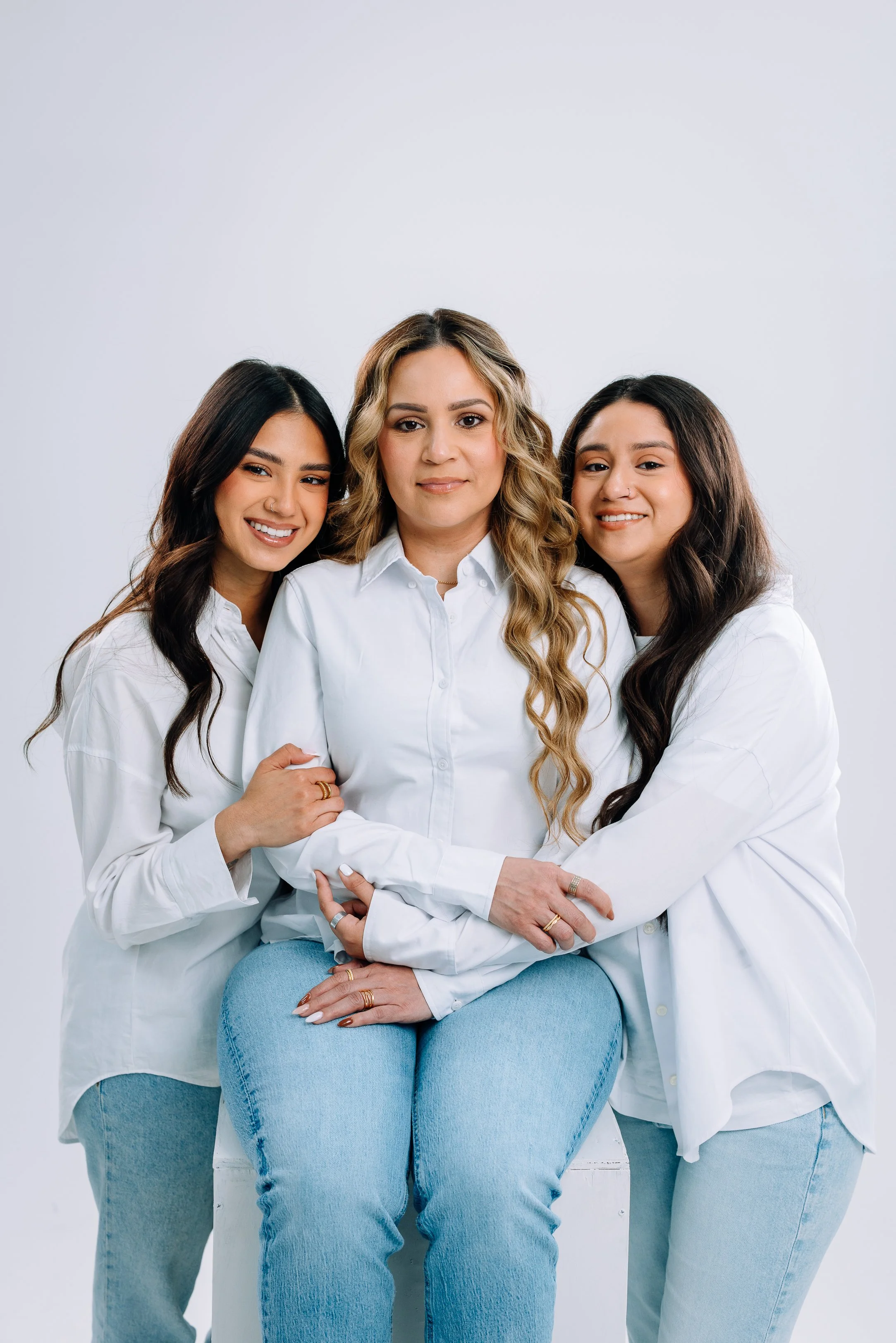 Three women wearing white shirts and light blue jeans embrace each other in a portrait against a plain white background.