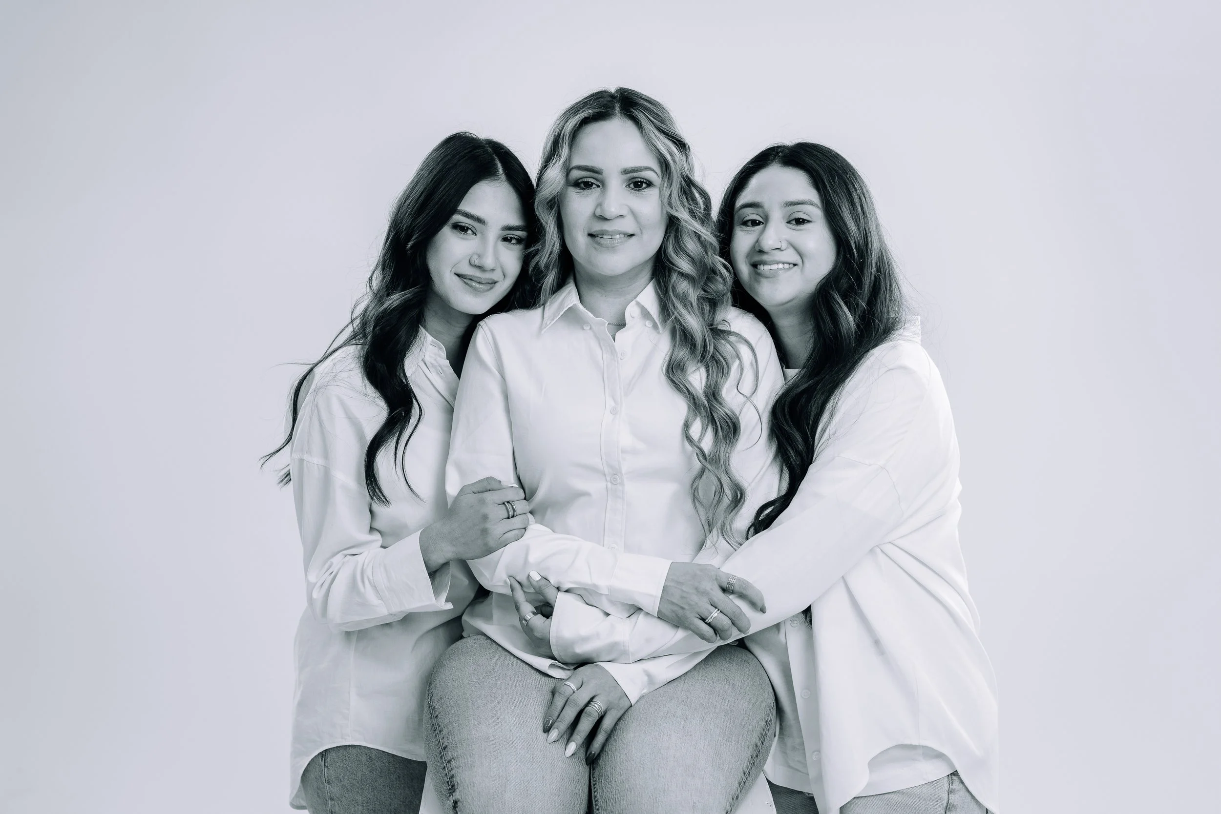 Three women, including a woman in the middle with blonde hair and two women with dark hair, dressed in white shirts, standing together against a plain background, looking at the camera.