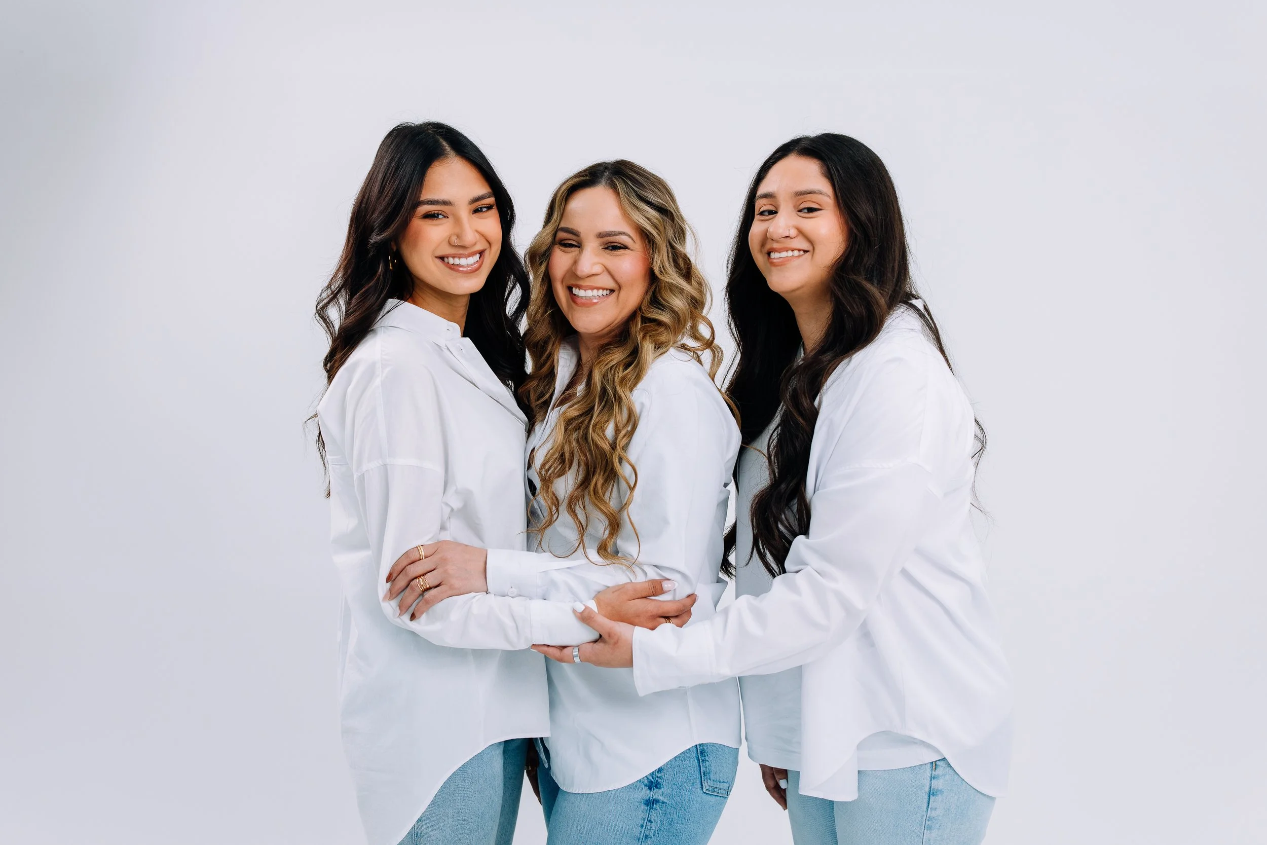 Three women smiling and hugging each other against a white background, wearing white shirts and blue jeans.