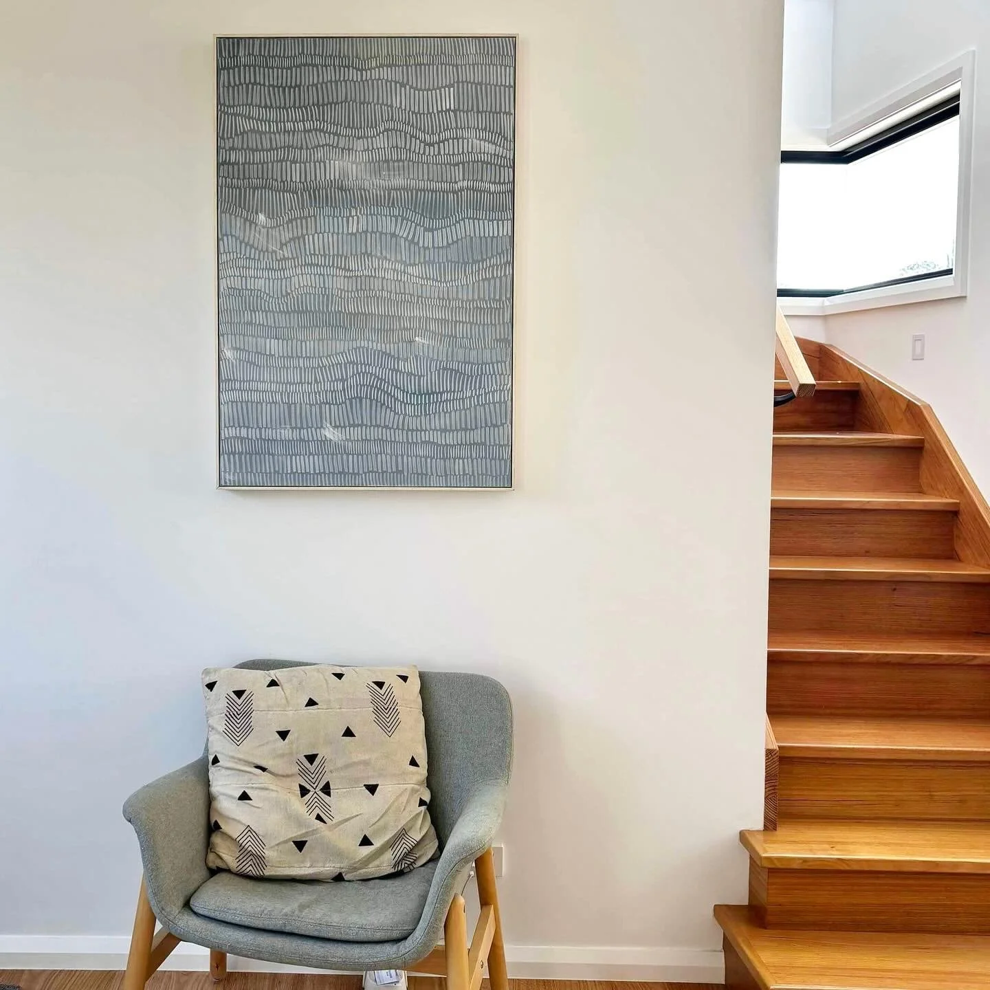 A cozy living space featuring a light gray armchair with a decorative black and white cushion, an abstract piece of wall art with a gray and black pattern hanging above, and a staircase with wooden steps leading upwards near a large window.