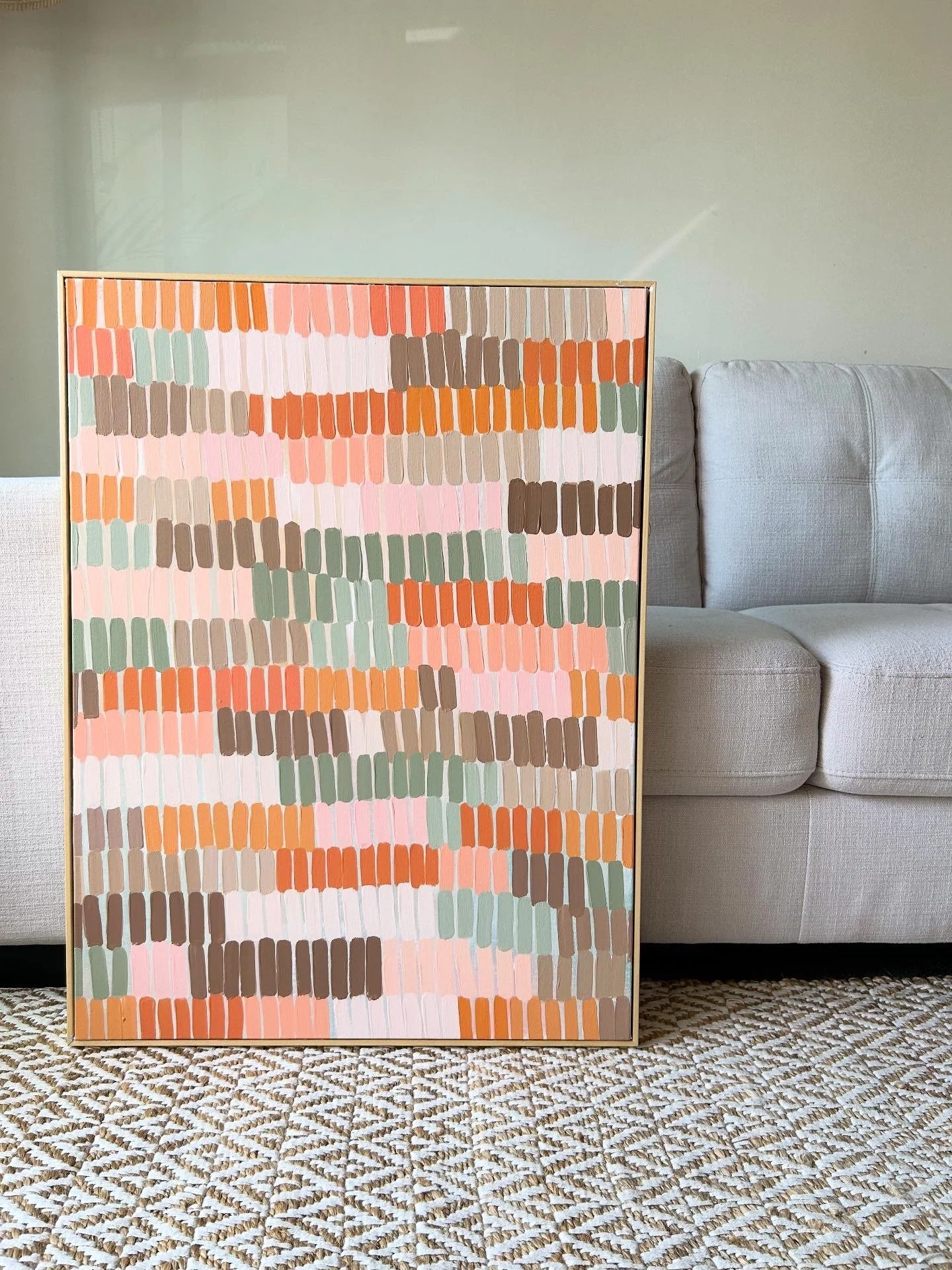 Colorful abstract painting with horizontal brushstrokes in shades of orange, pink, brown, green, and white, leaning against a beige sofa in a room with a patterned rug.