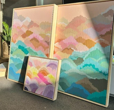 Three colorful abstract paintings of mountain-like shapes in pastel colors, displayed on a table with natural light.