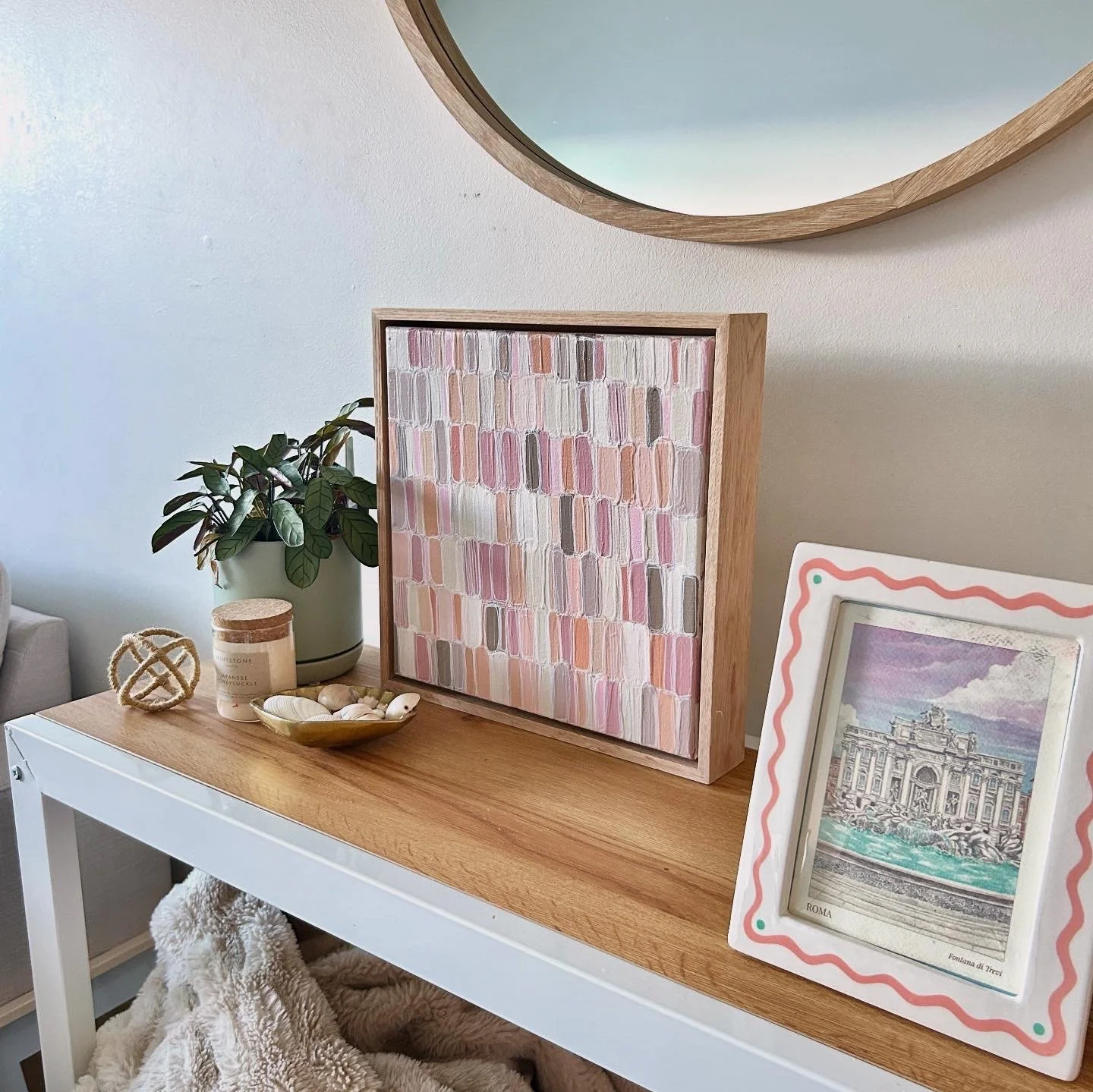 Decorative table with a green potted plant, a framed colorful abstract artwork, a framed vintage-style print of Rome, a candle, and shells, against a beige wall with a round mirror.