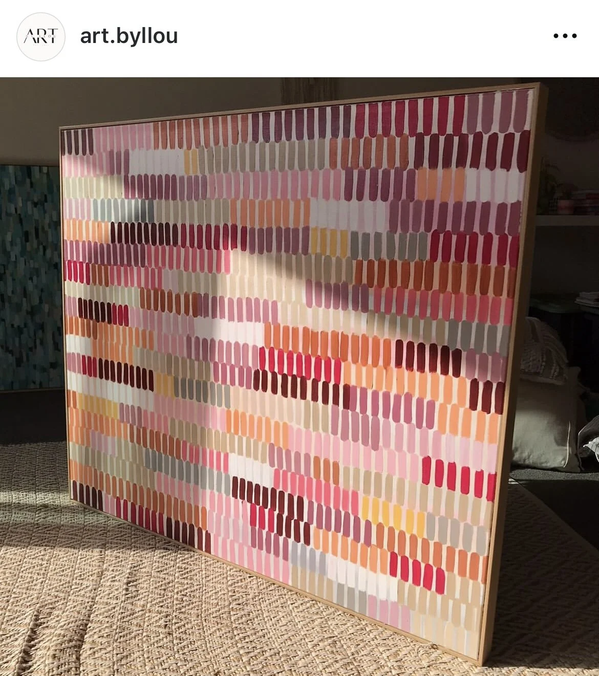 Abstract mixed media artwork with colorful rectangular brushstrokes in pink, red, orange, beige, gray, and white on a large canvas, leaning against a wall in a room with sunlight.