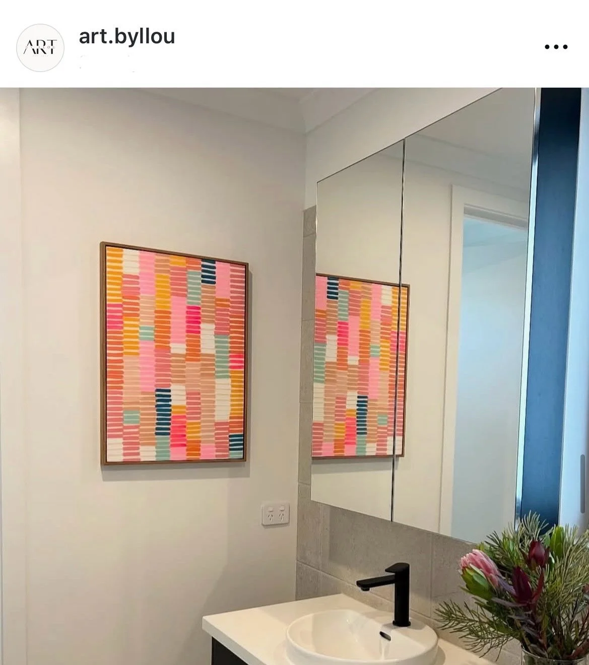 A bathroom vanity with a white sink, black faucet, and a mirror cabinet. Two colorful, geometric framed artworks hang on the wall. A small pink flower arrangement is on the right side of the sink.