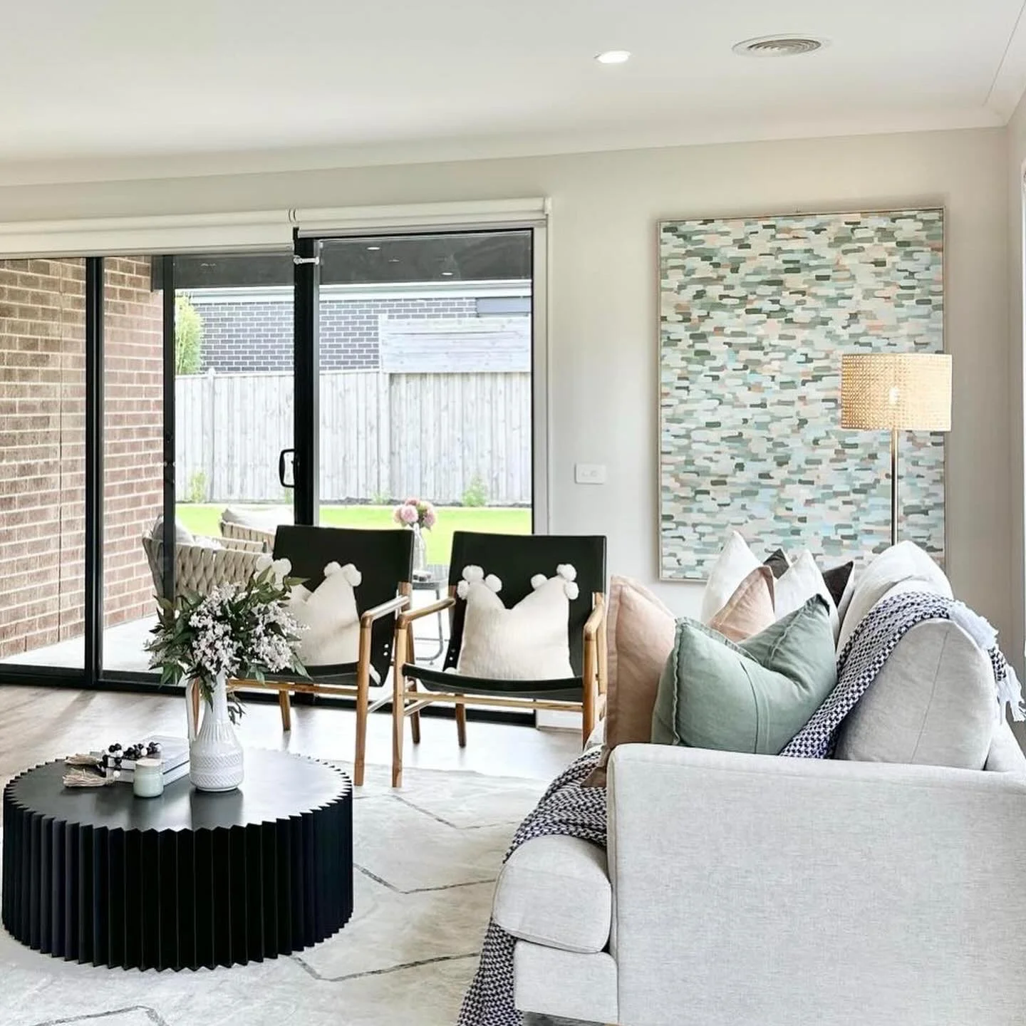 Bright living room with large sliding glass doors leading to backyard, contemporary artwork on wall, white sofa with pastel pillows, black round coffee table with vase of flowers, armchairs, and floor lamp.