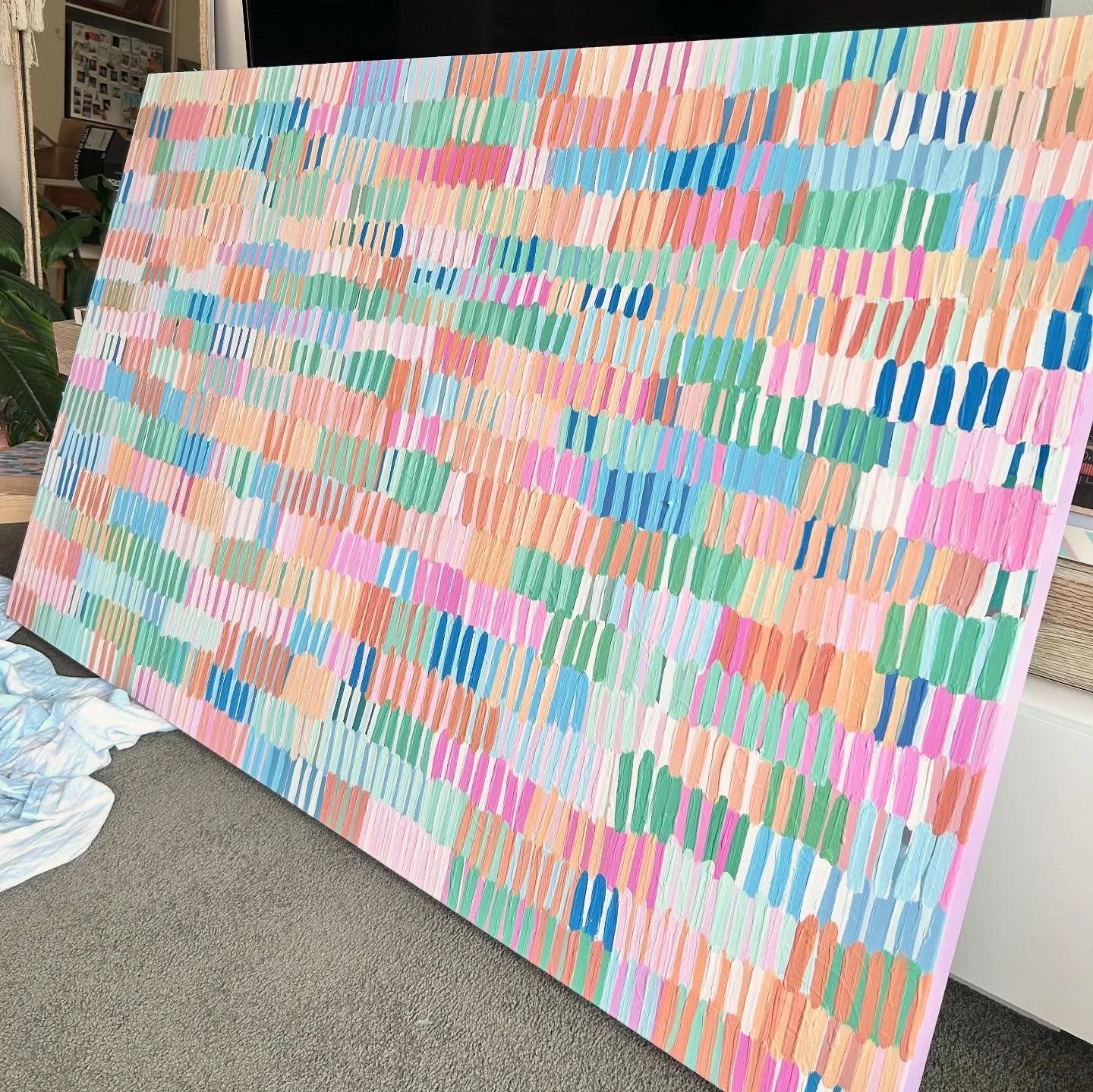 Large colorful abstract painting with horizontal brushstrokes in shades of pink, orange, blue, green, and white, leaning against a wall in a room with a carpeted floor.