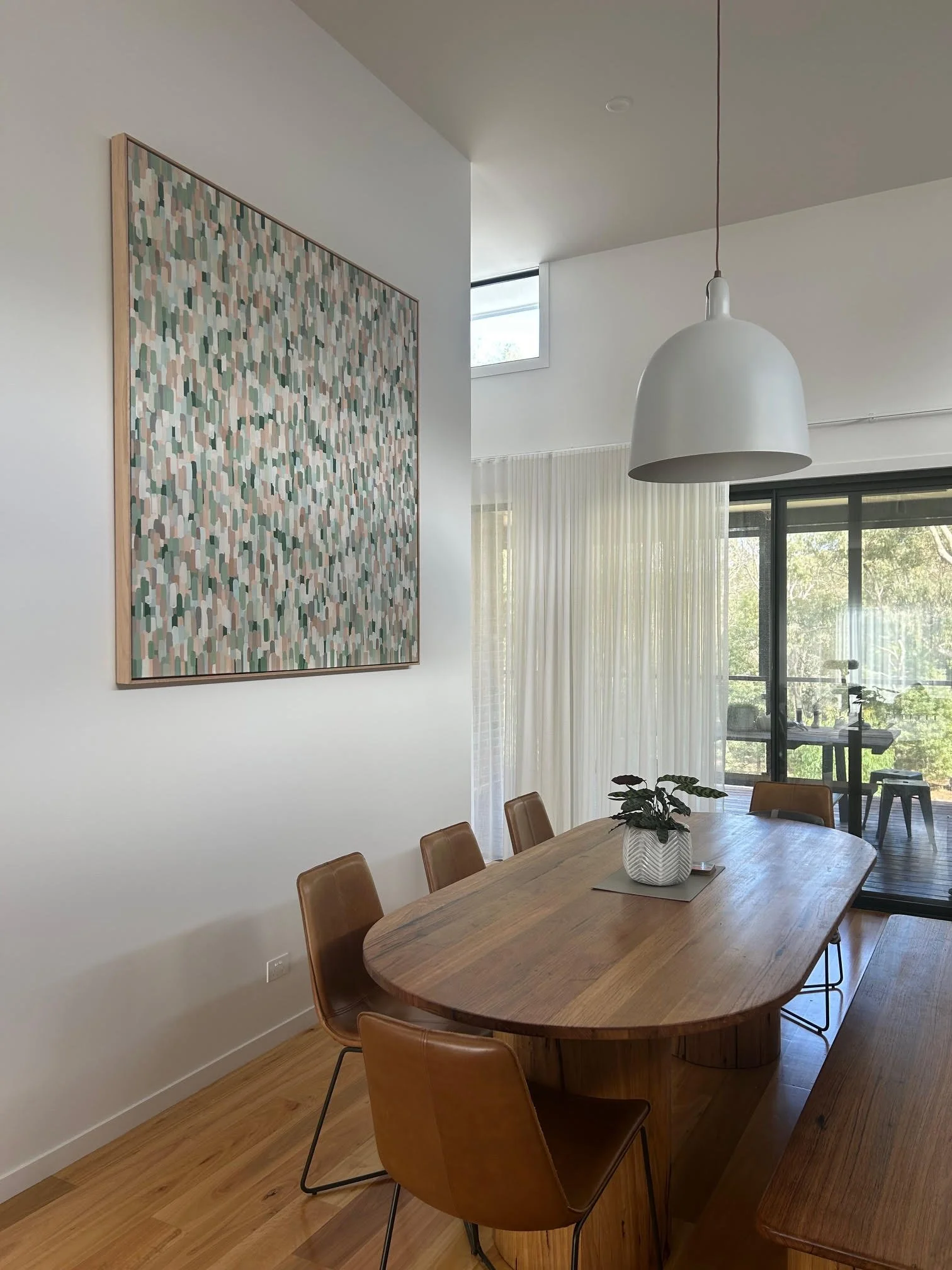 Modern dining room with wooden table and tan chairs, large abstract wall art, white pendant light, glass doors leading to balcony with exercise bike and greenery outside.