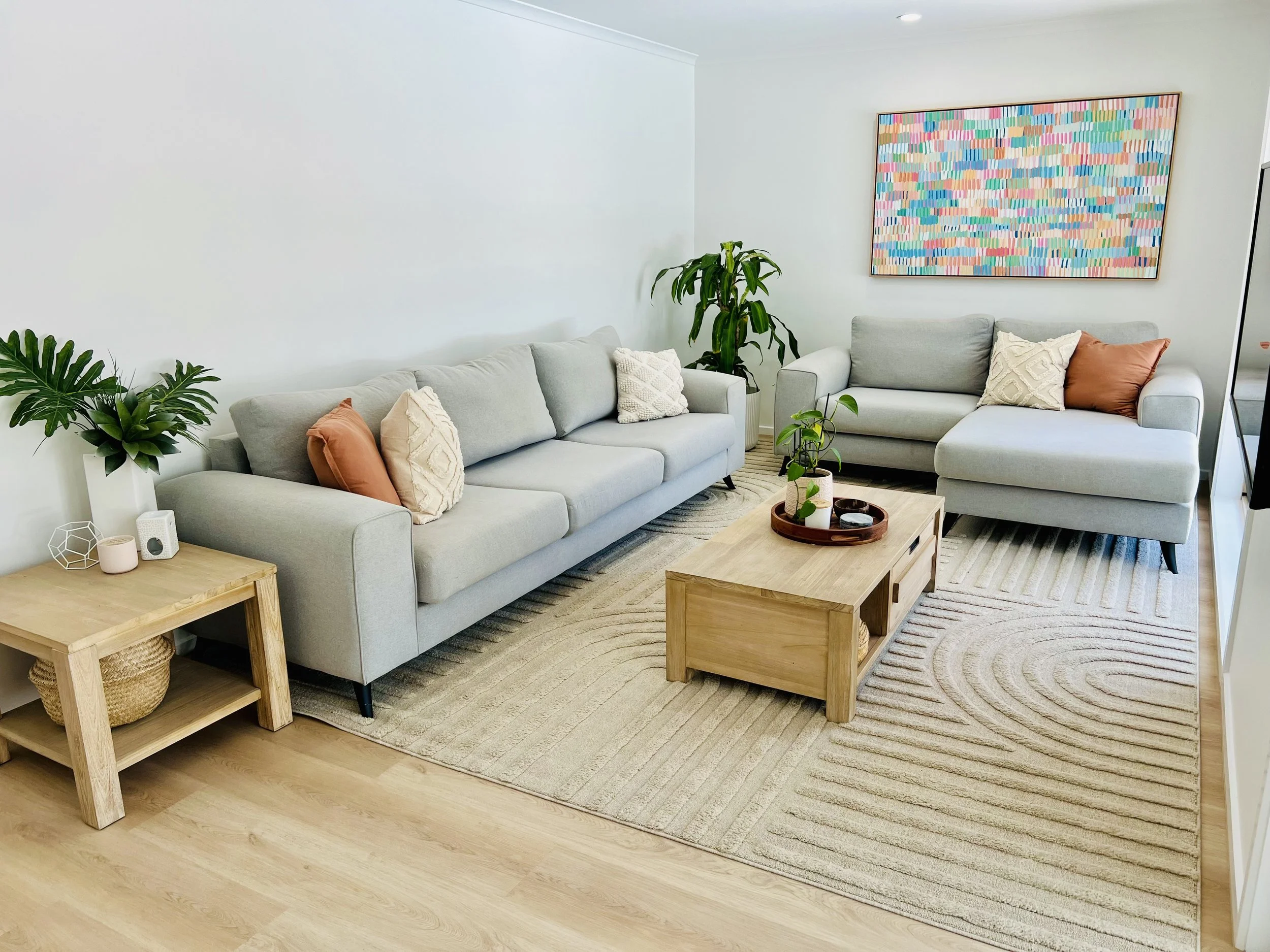 A modern living room with two light gray sofas decorated with patterned and solid-colored pillows, a wooden coffee table with plants and small decor items, a beige rug with a circular pattern, a side table with a plant and decorative objects, and abs