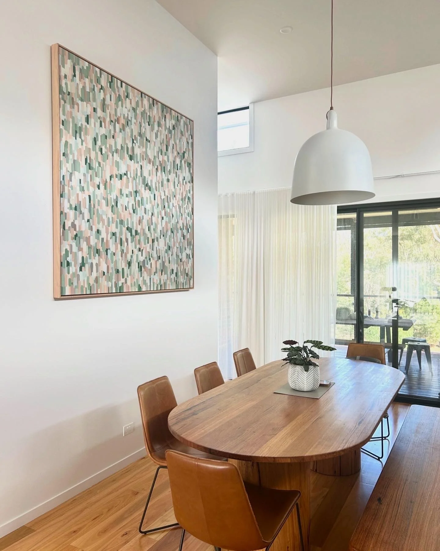 A dining room with a wooden table surrounded by leather chairs, a plant on the table, and a large abstract painting on the wall. There are large windows with curtains and a balcony outside.