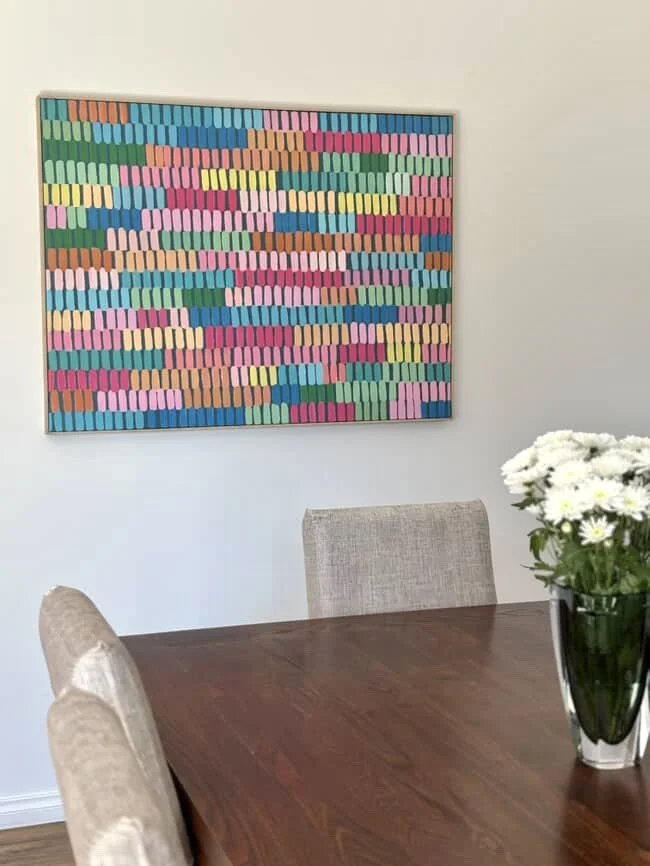 Colorful abstract artwork hanging on a white wall in a dining room with a wooden table, beige chairs, and a vase of white flowers.