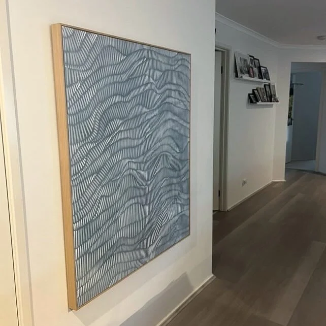 A framed abstract art piece featuring wavy blue and white lines hanging on a white wall in a home interior.