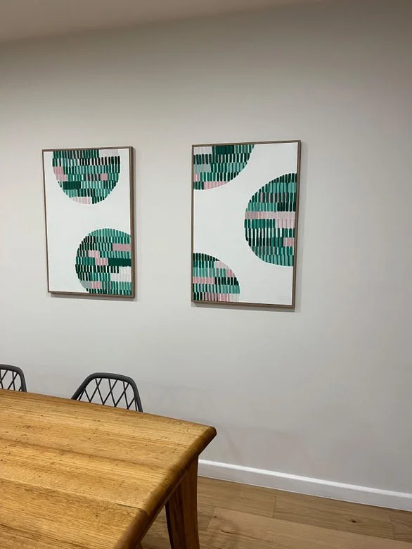 Two abstract framed artworks with geometric shapes and green, pink, and black stripes on a white wall