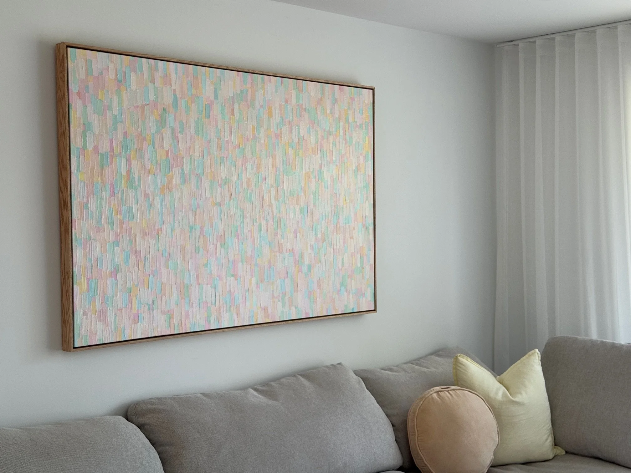 Living room with a pastel-colored abstract painting on the wall above a beige sectional sofa with yellow and light pink pillows, next to white curtains.