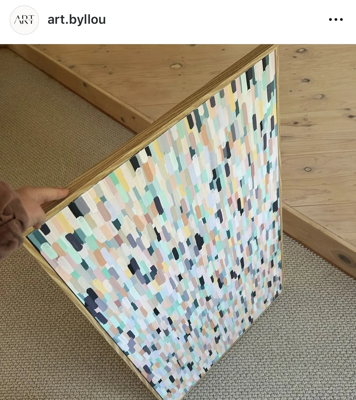 A hand holding a colorful abstract painting with rectangular brushstrokes, leaning against a wooden wall.