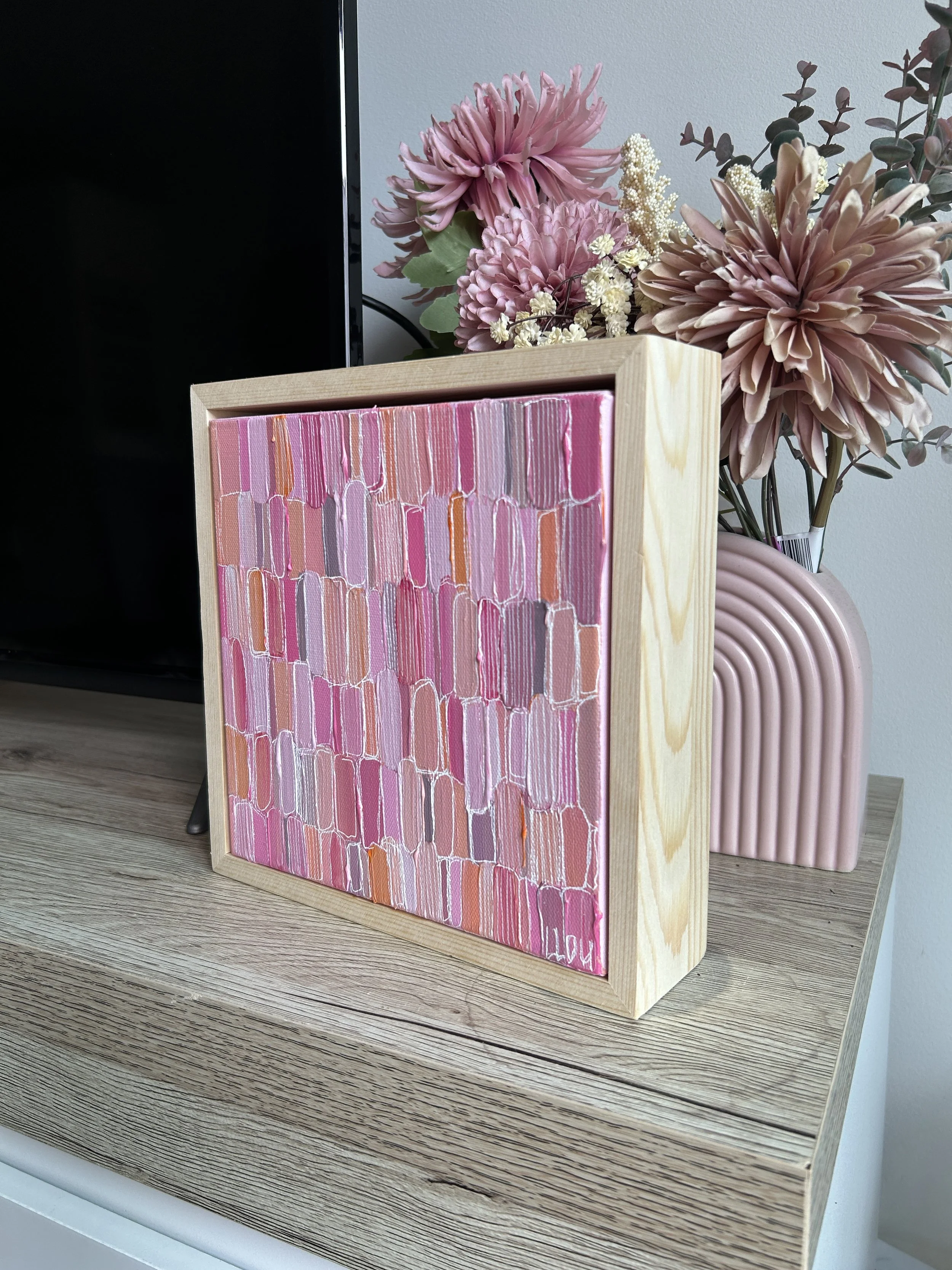 A small framed abstract painting with pink and purple brushstrokes, placed on a wooden surface beside a pink ribbed ceramic vase with pink flowers.