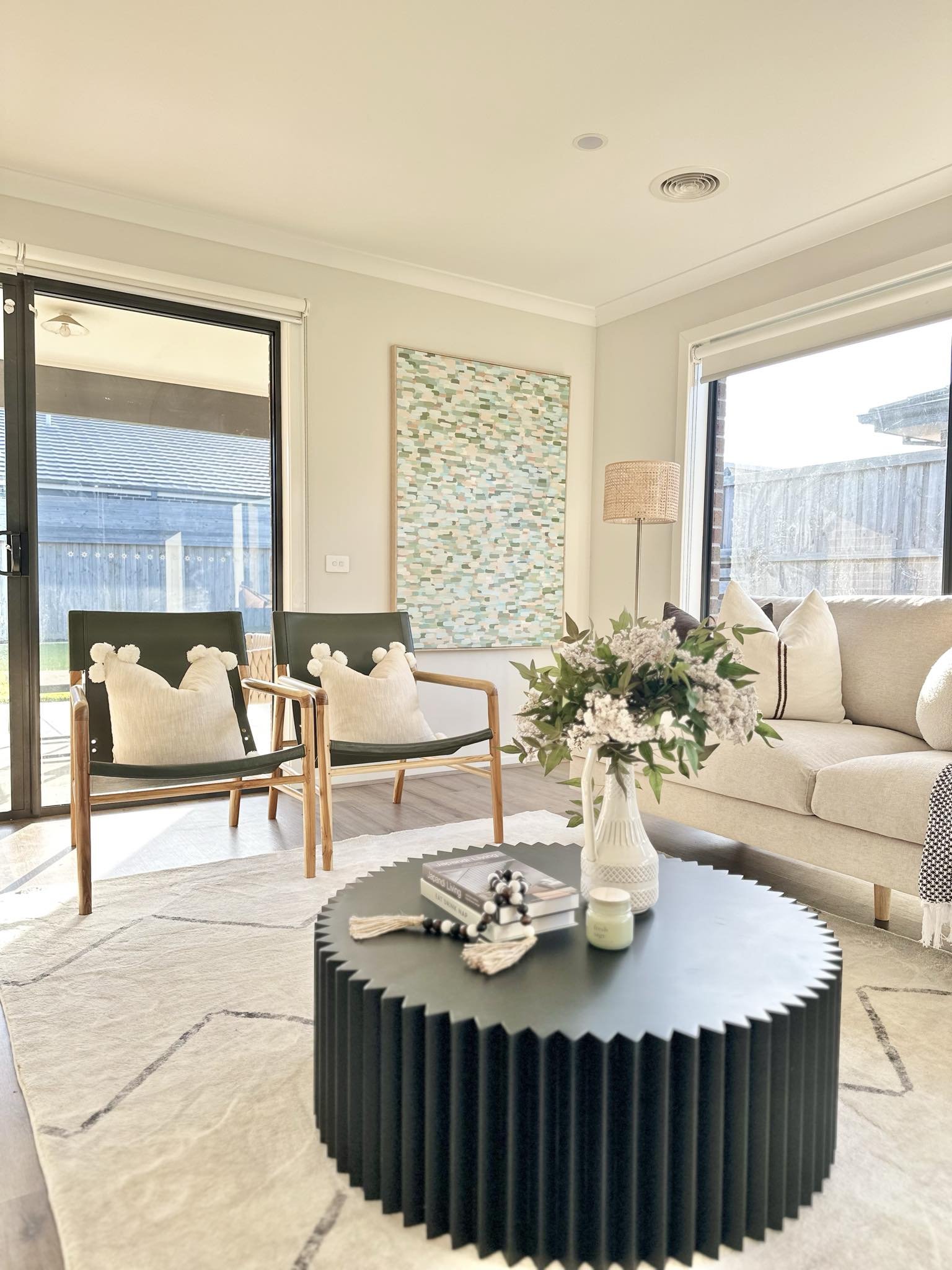 Bright living room with beige sofa, two black and wood chairs with white cushions, a black circular coffee table with a vase of flowers, books, a candle, and some decor, large windows and sliding glass door letting in natural light, artwork on the wa