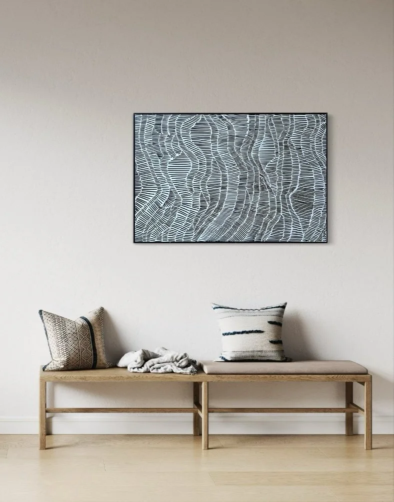 A minimalist wooden bench with three decorative pillows and a blanket against a plain white wall with abstract black and white artwork.