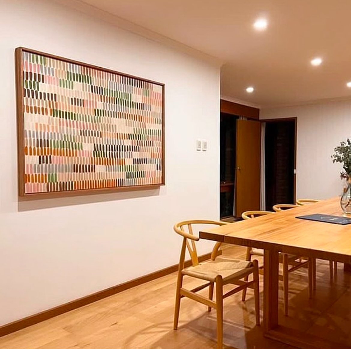 A dining room with wooden flooring, a large wooden table, and matching chairs. There is a colorful abstract art piece on the wall and a vase with flowers on the table. The room has recessed ceiling lights and an open door leading to another space.