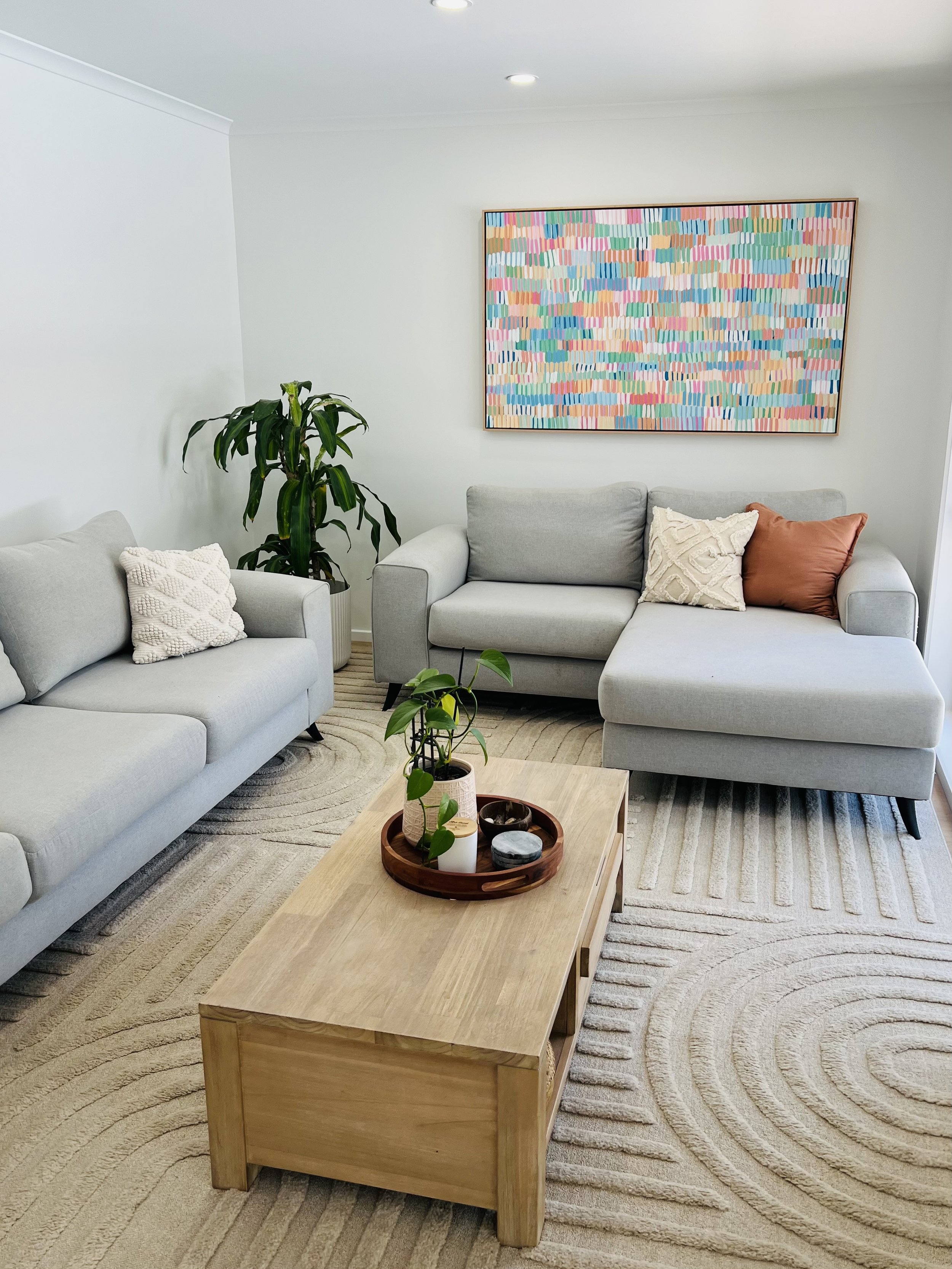 A modern living room with two light gray sofas, decorative pillows, a wooden coffee table with a tray holding a potted plant and candles, a large colorful abstract painting on white walls, a tall indoor plant, a beige rug with circular patterns, and 