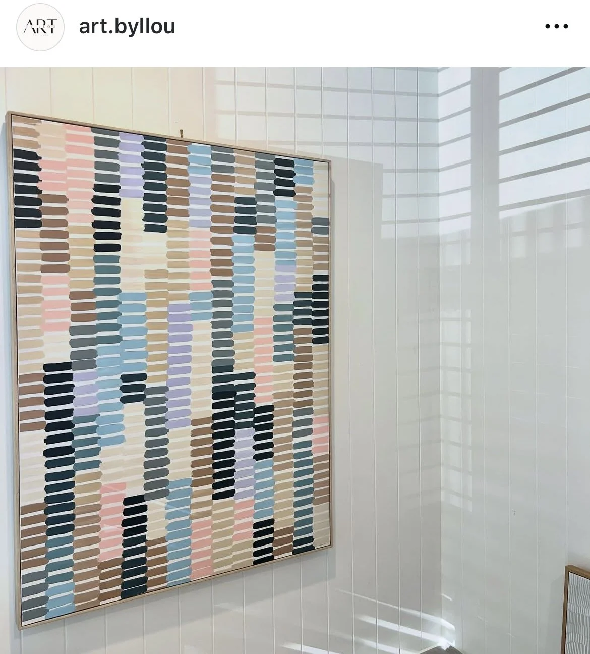 A colorful abstract painting with horizontal brushstrokes in shades of pink, purple, blue, black, beige, and brown, framed and hung on a white wall with sunlight and window shadows.