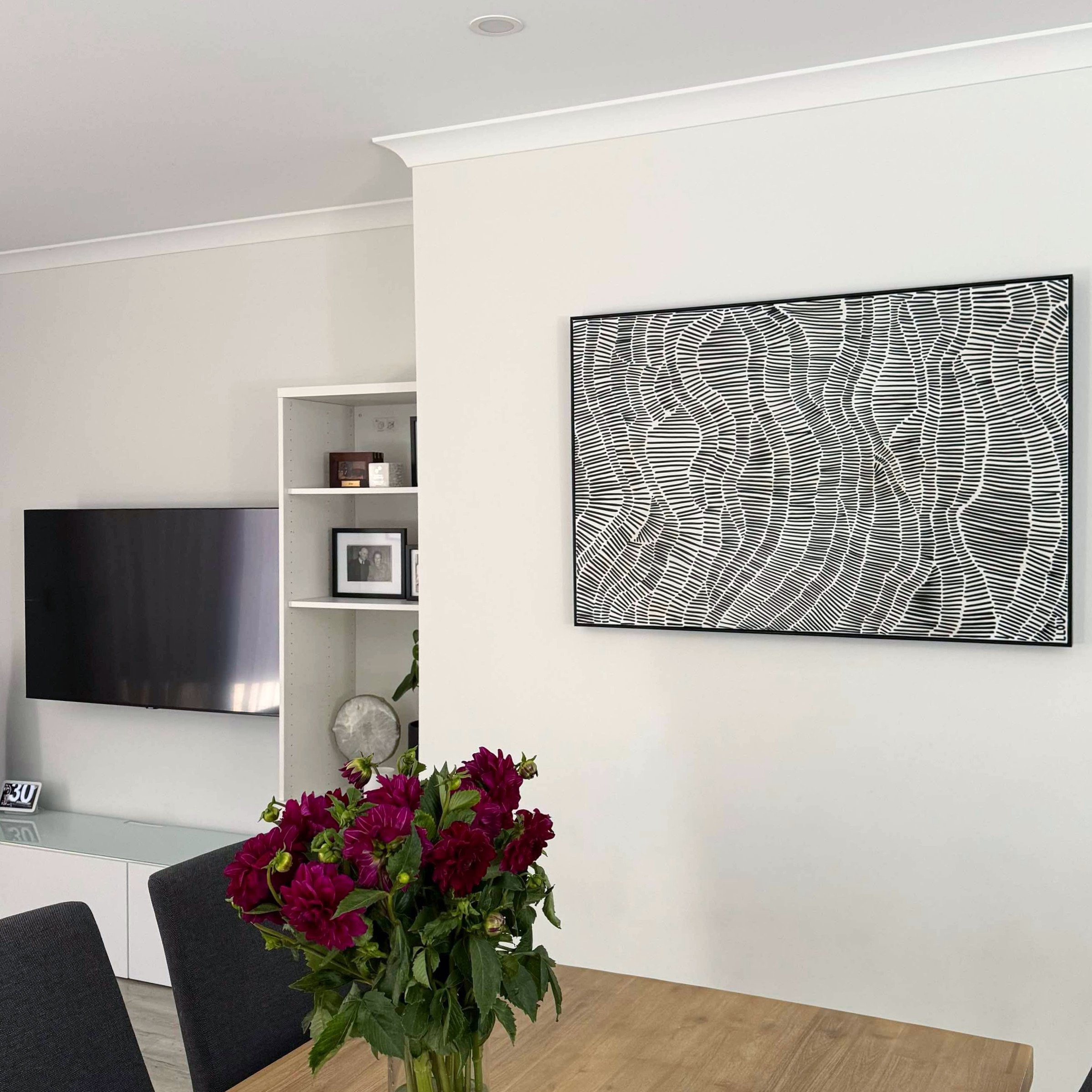 Modern living room with white walls, large wall art with black and white abstract pattern, a mounted flat-screen TV, a white shelving unit with framed photos and decor, a dining table with dark chairs, and a vase of dark pink flowers on the table.