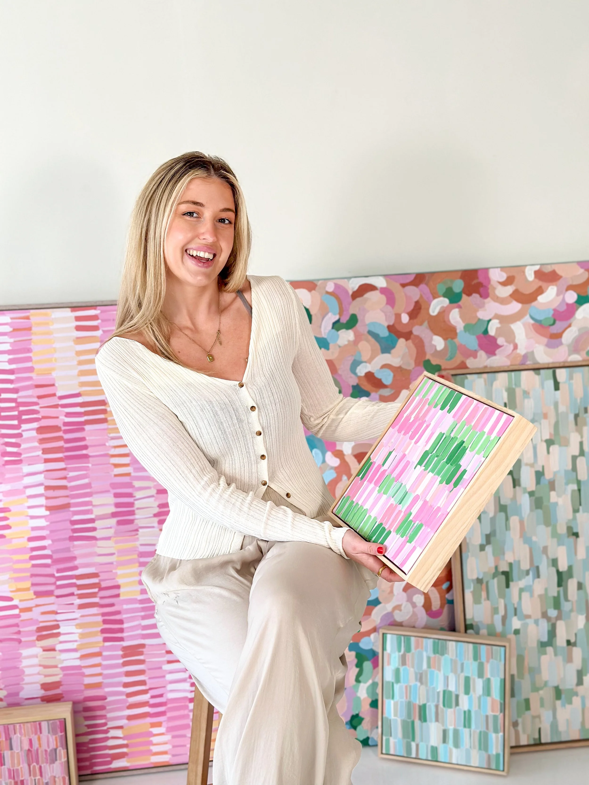 A woman with long blonde hair smiles while sitting on a stool, holding a framed abstract artwork with pink and green brushstrokes. Behind her are colorful patterned canvases leaning against a wall.