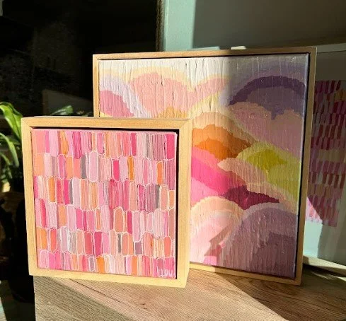 Two abstract colorful paintings with pink, purple, yellow, and orange brushstrokes on wooden frames, displayed on a wooden surface.