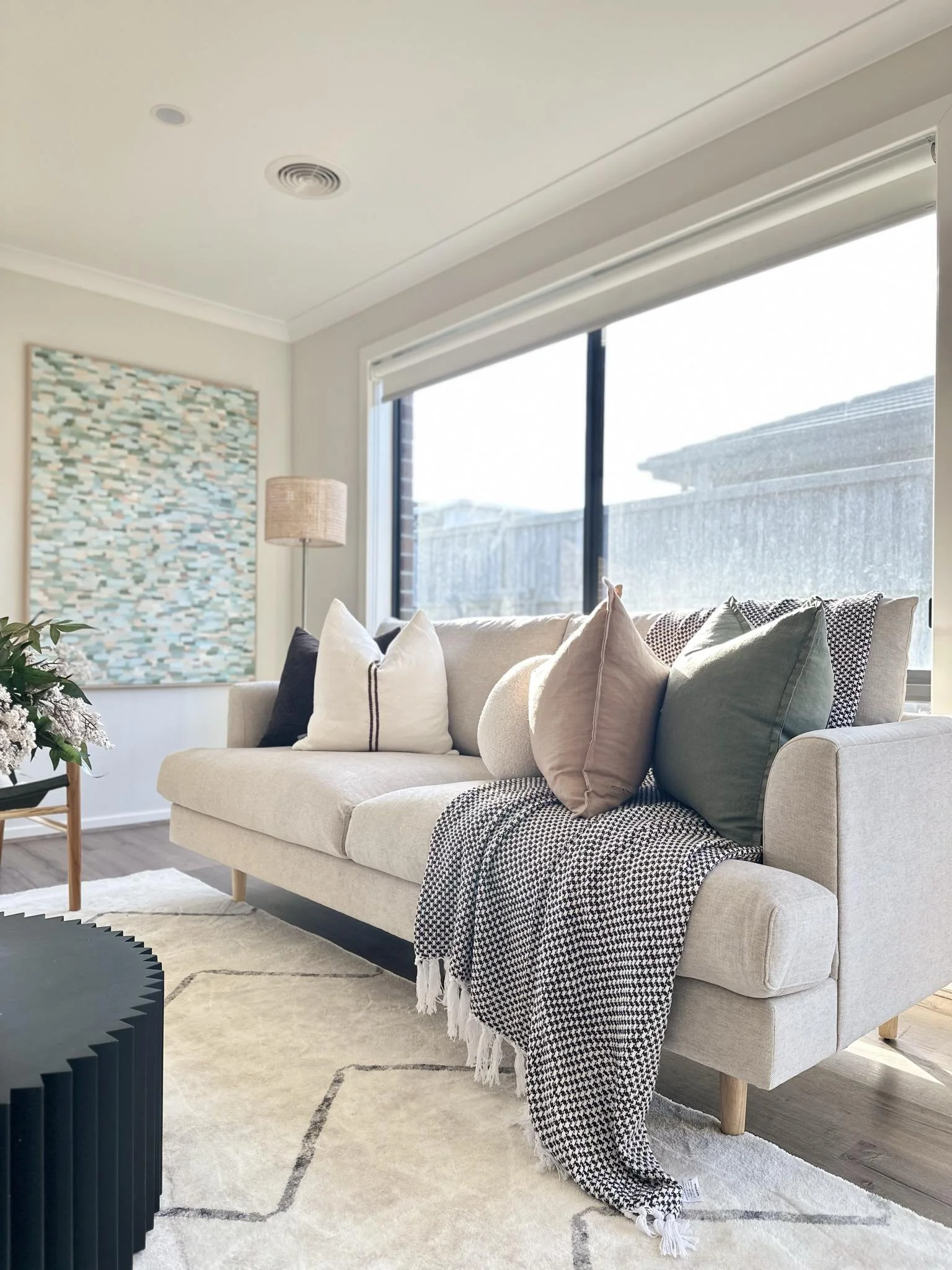 Living room with beige sofa, throw pillows, patterned blanket, large window, artwork, lamp, and decorative items.
