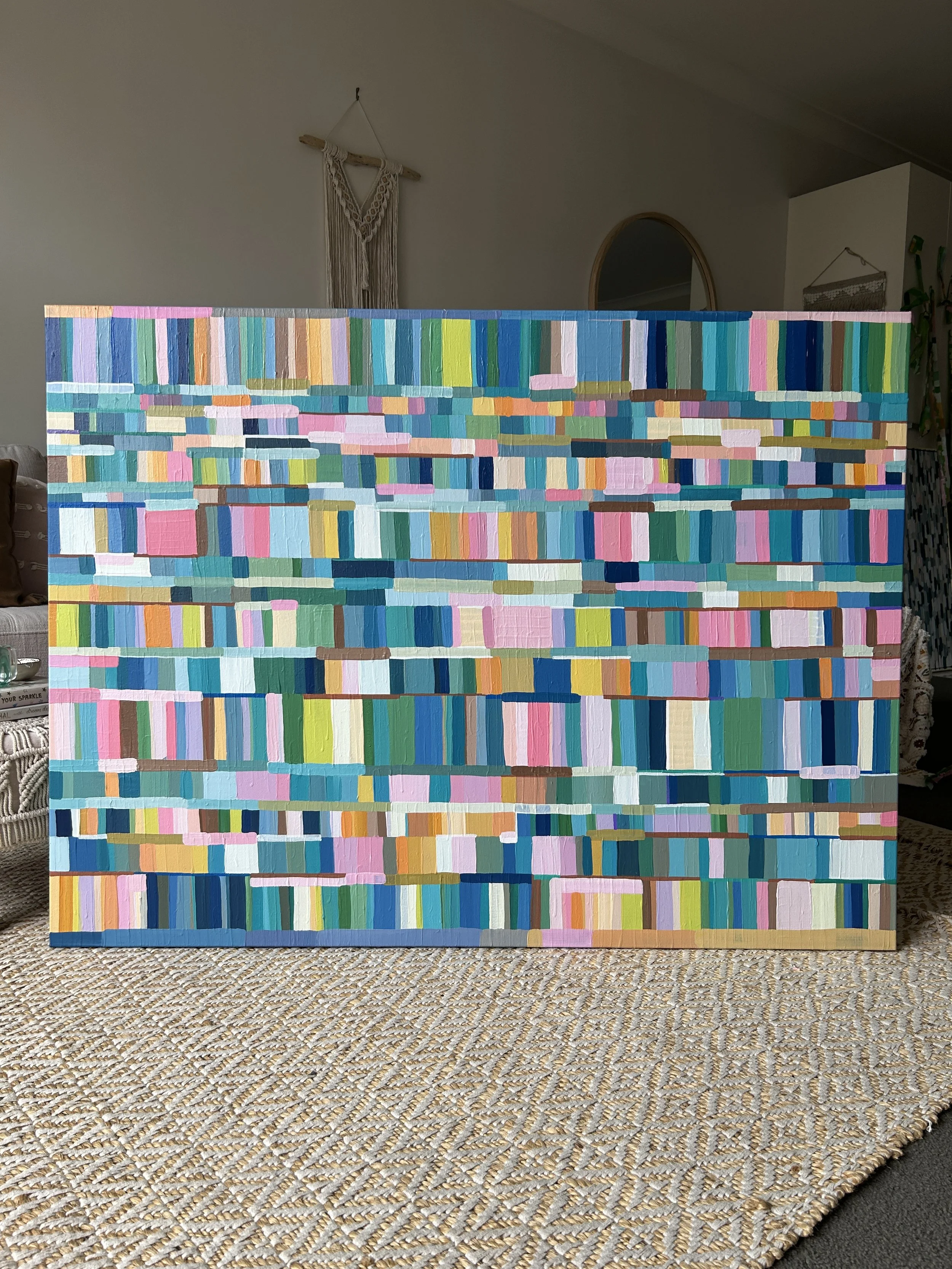 Abstract colorful painting with horizontal rectangles of various shades of blue, pink, green, yellow, and orange, displayed indoors on a woven table surface.