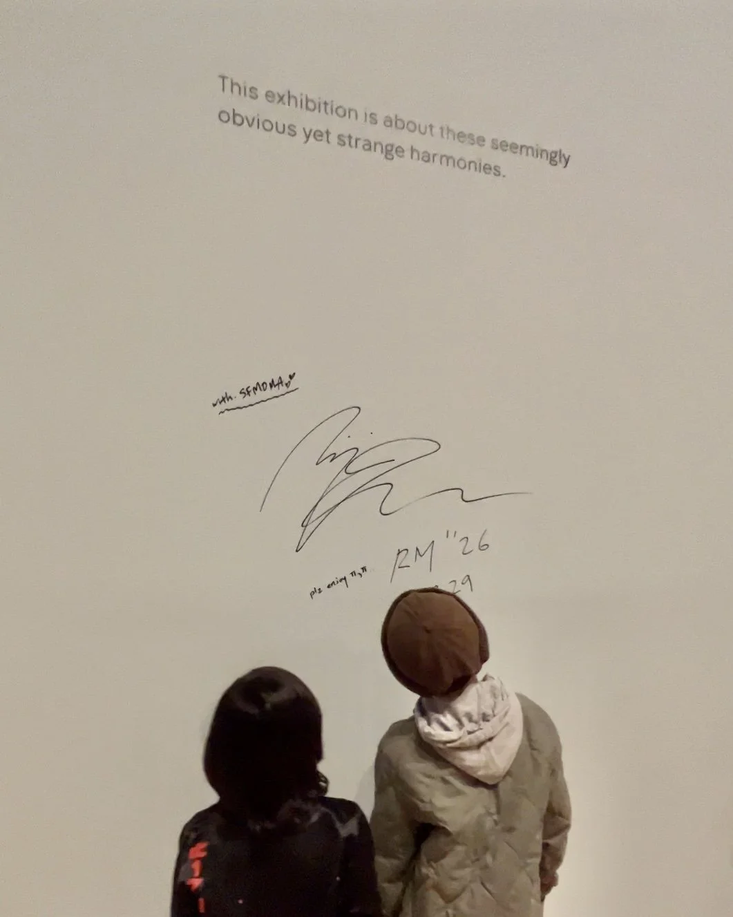 Two visitors stand beneath RM's handwritten message and signature on a gallery wall at RM x SFMOMA: Between You and Me.