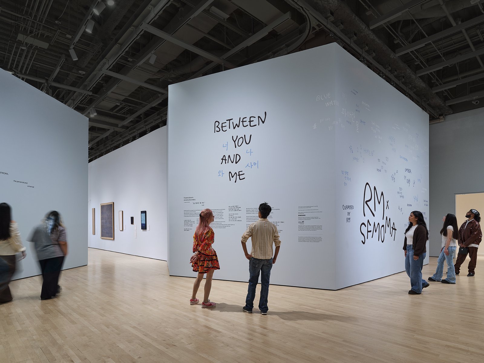 Entrance installation at RM x SFMOMA: Between You and Me, featuring large handwritten exhibition lettering and visitors exploring the seventh-floor galleries at the San Francisco Museum of Modern Art.