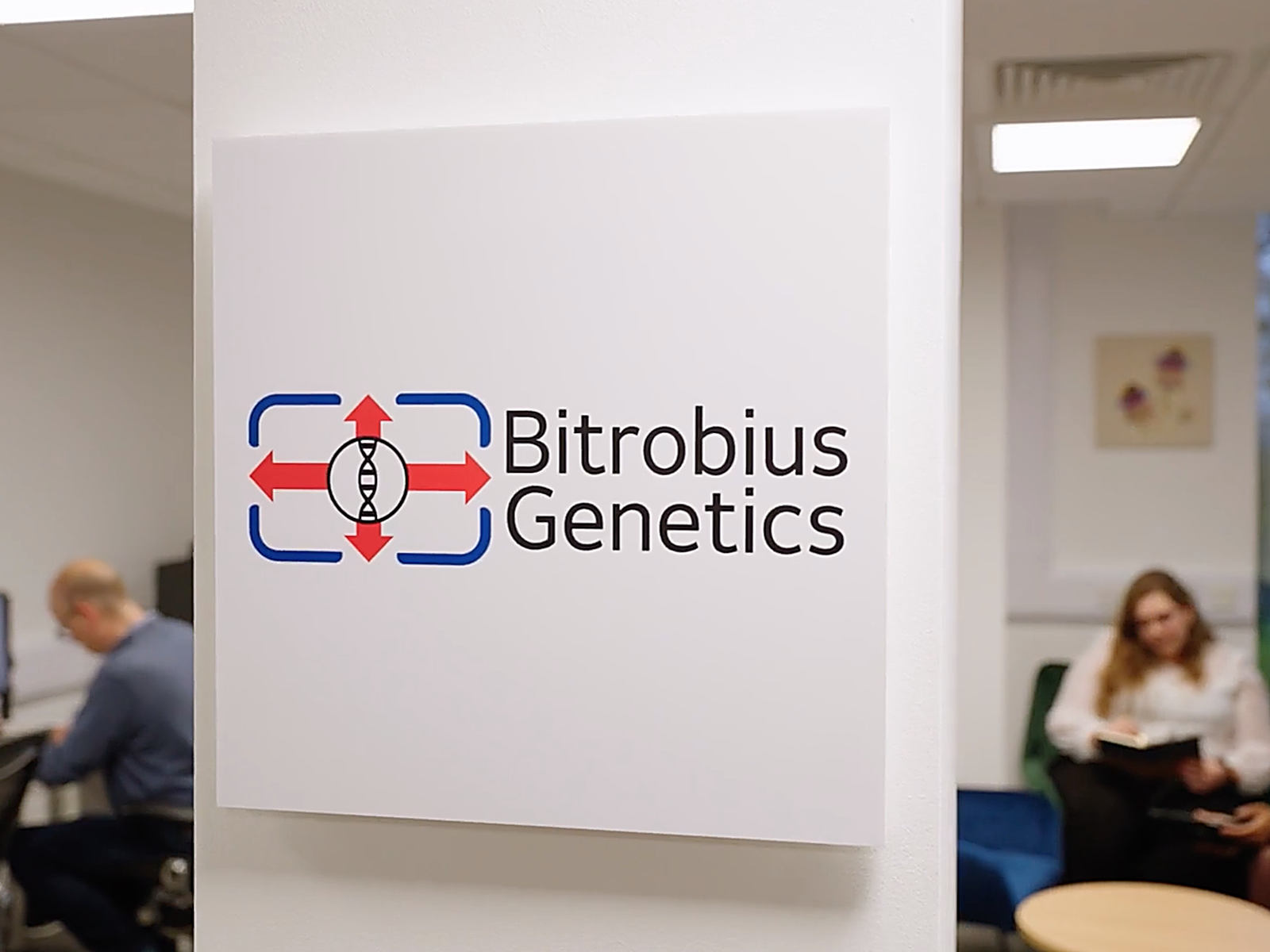 Sign for Bitrobus Genetics on a white wall at an office, with two people working in the background.