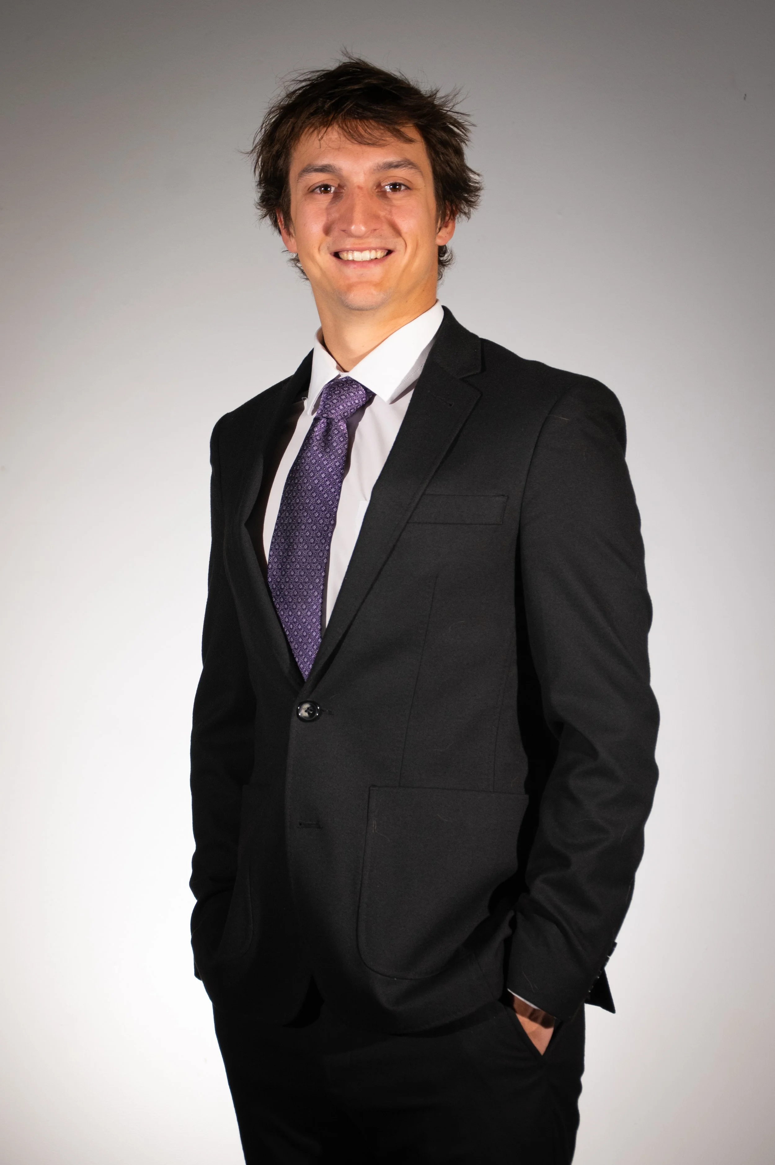 Man in a black suit with a purple tie standing against a light background, smiling.