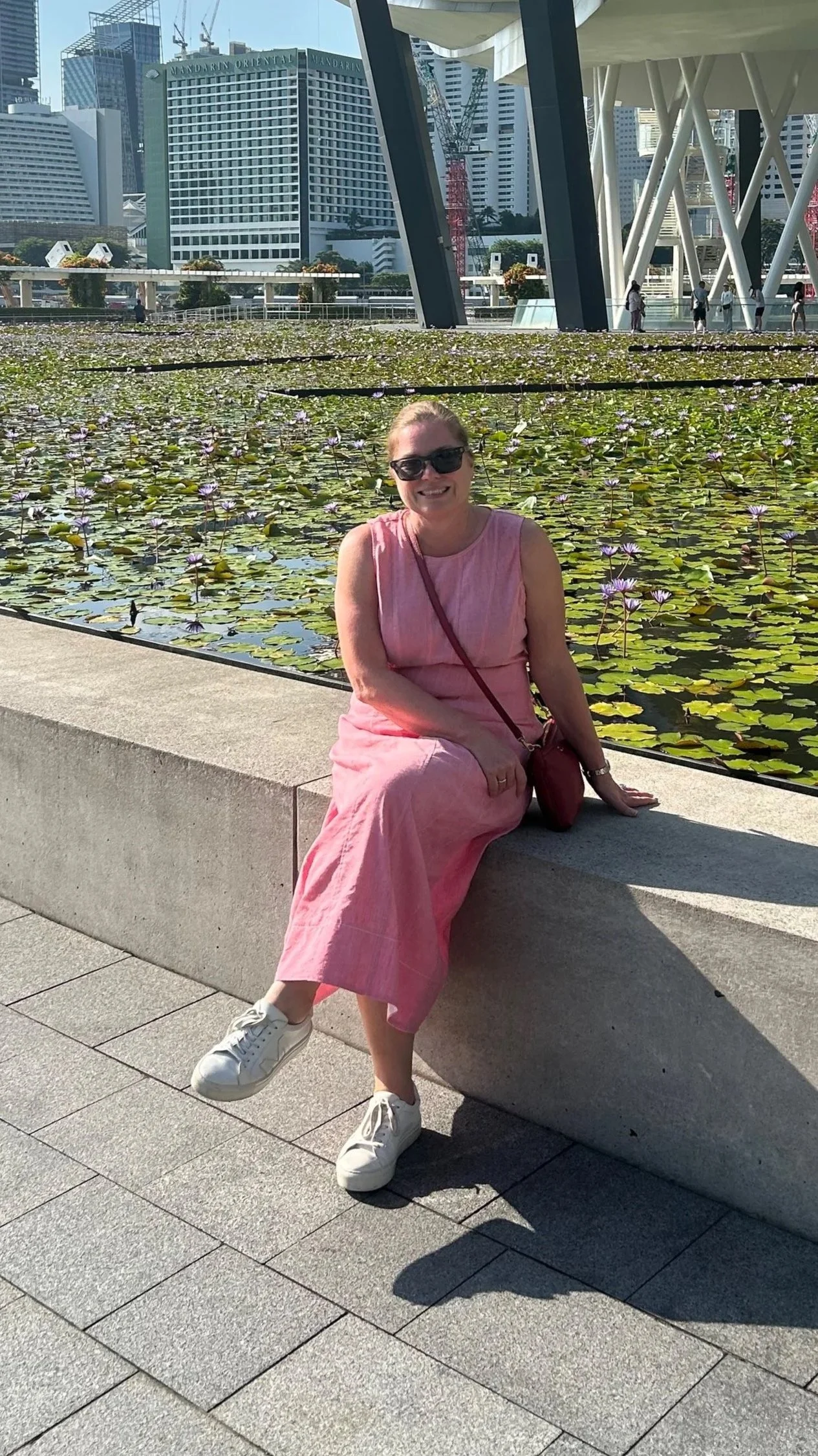 Woman sitting on a concrete bench near a pond filled with water lilies in an urban park, with modern city skyscrapers in the background.