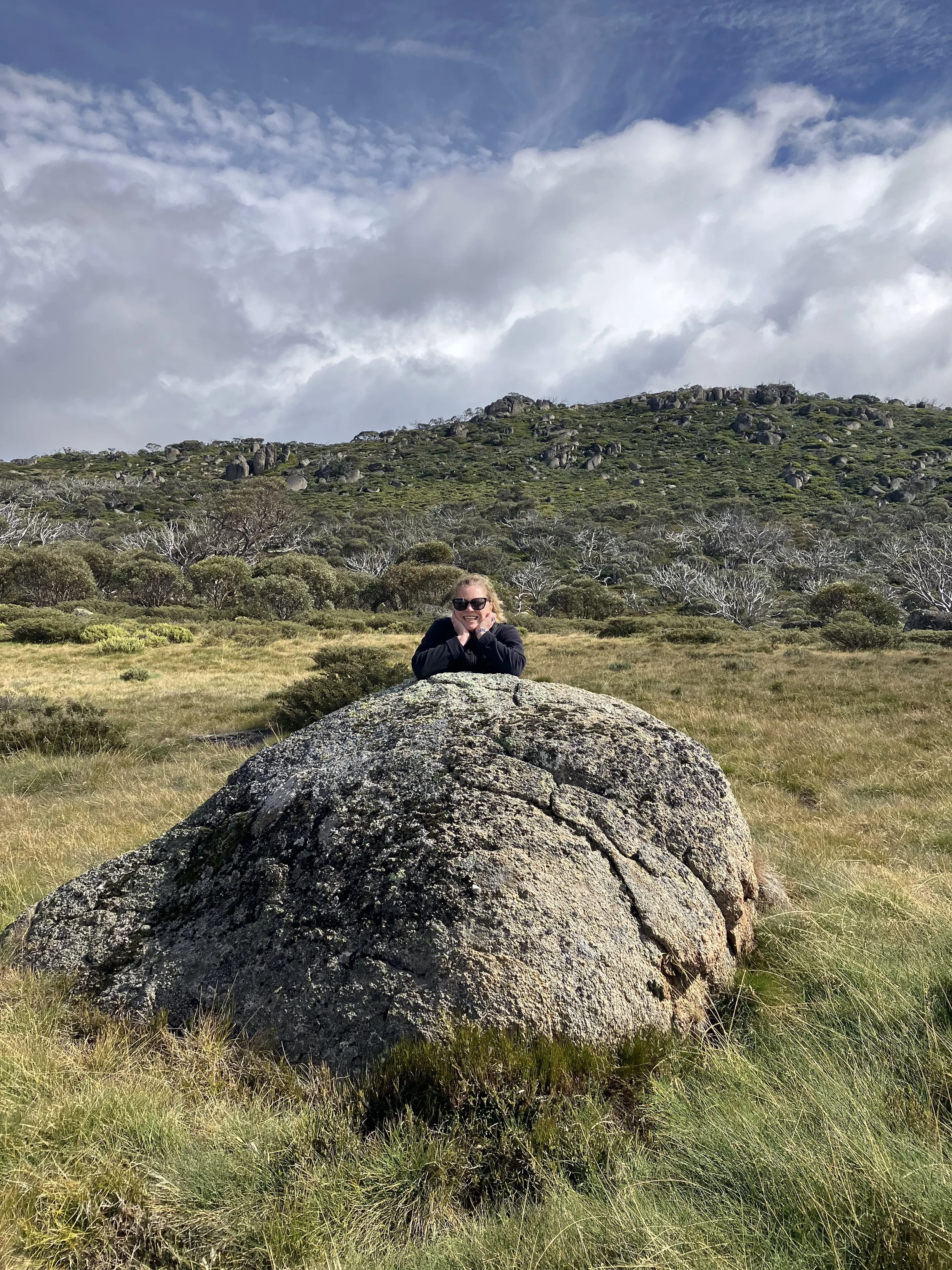 A woman with sunglasses smiling and resting her chin on her hands, leaning on a large moss-covered rock in an open grassy landscape with trees and a rocky hill under a cloudy sky.